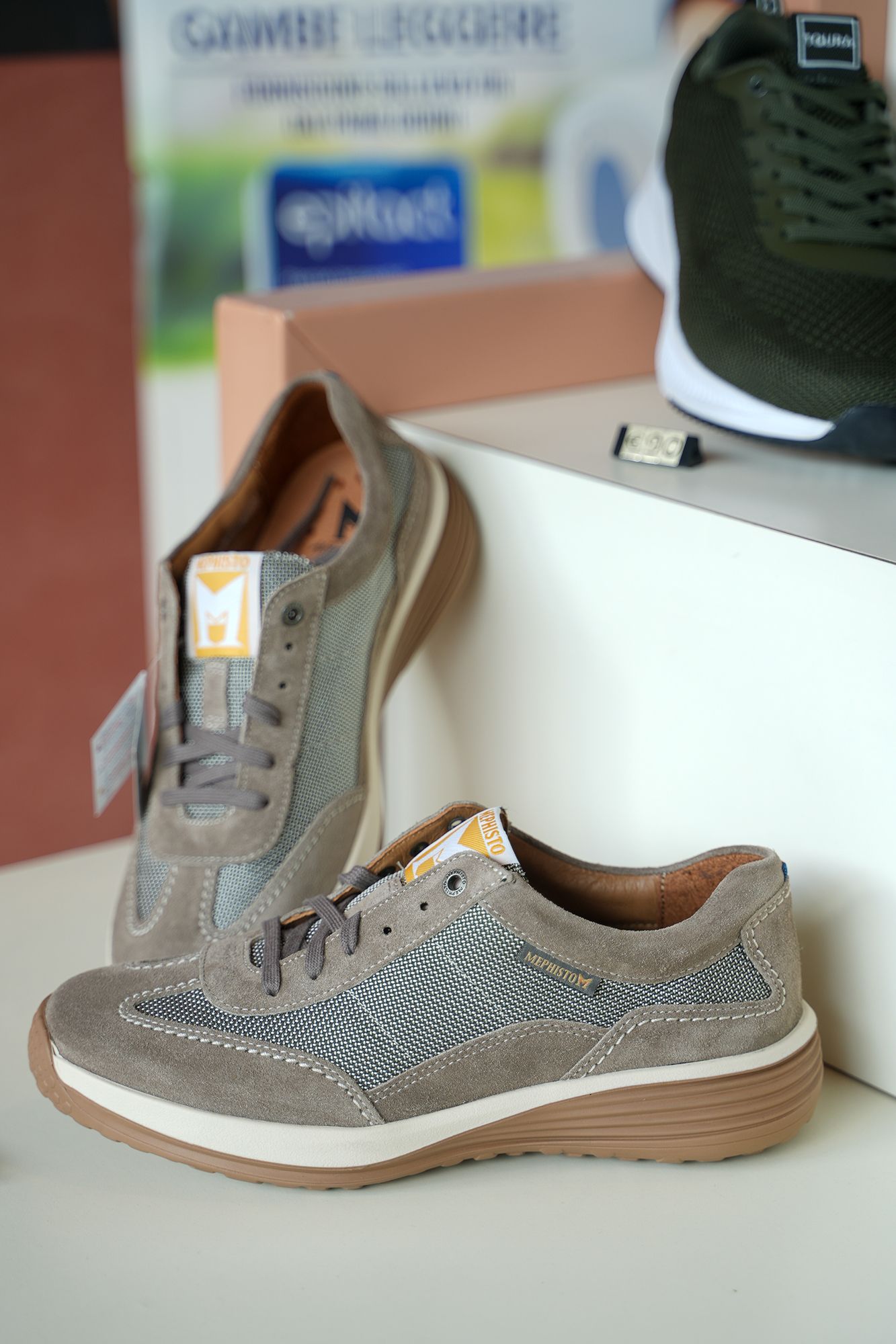 Due scarpe da ginnastica color cuoio con dettagli in rete su una superficie bianca, un'altra scarpa sullo sfondo.