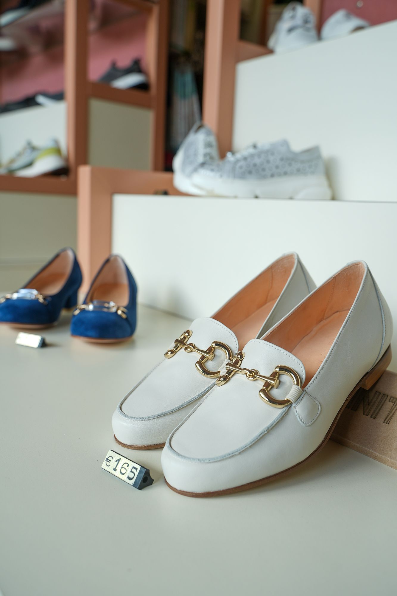 Mocassini bianchi con dettagli dorati, esposti insieme ad altre scarpe sugli scaffali di un negozio.