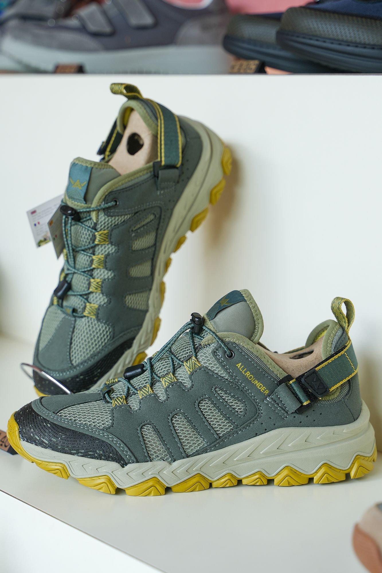 Scarpe da trekking grigie e gialle esposte su uno scaffale bianco.