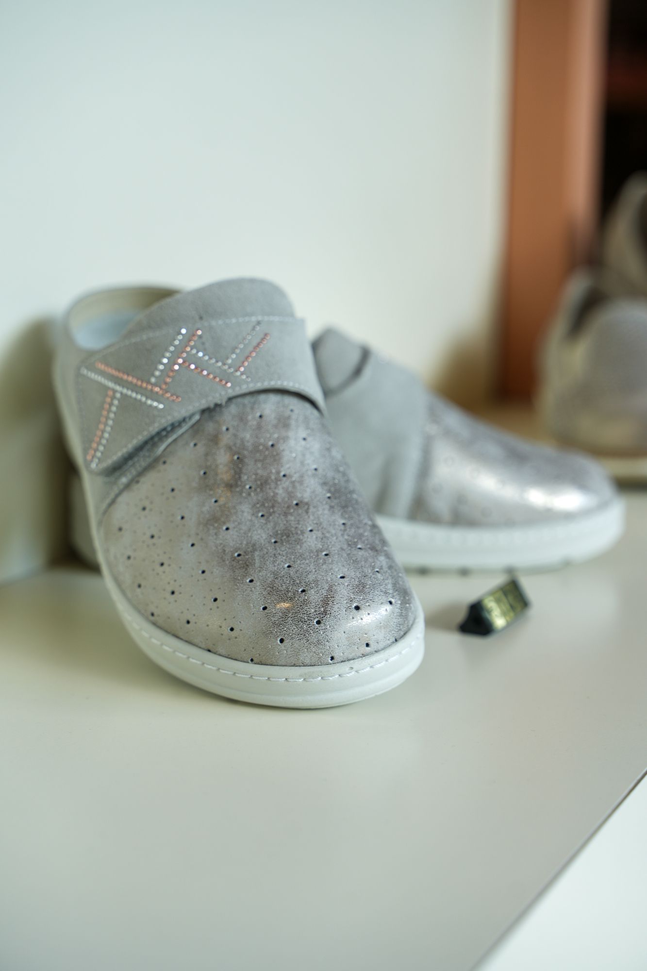 Scarpe slip-on argentate con dettagli glitterati e cinturino, esposte su uno scaffale bianco.