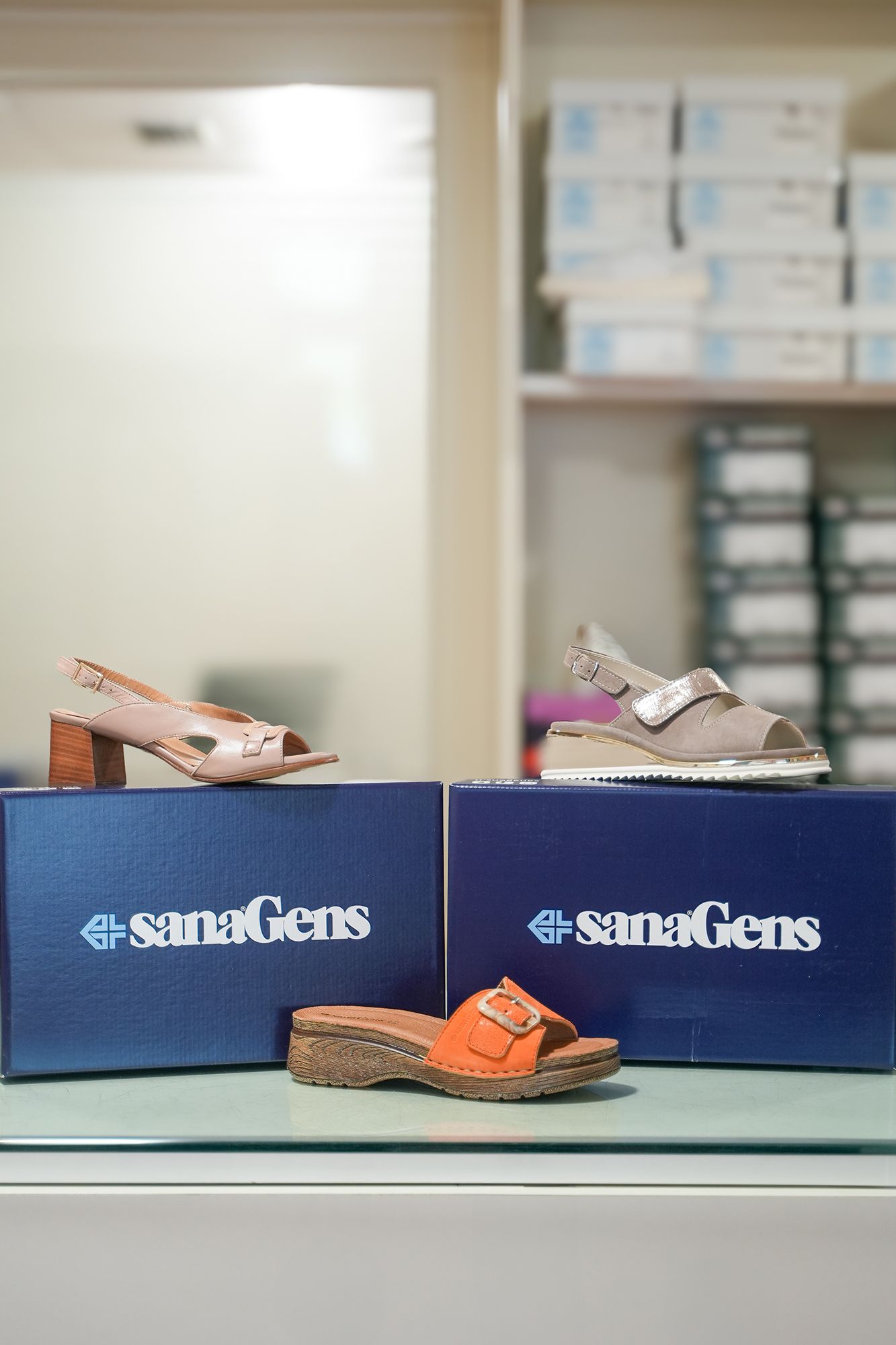 Tre paia di sandali SanaGens in scatole blu, esposti su un bancone con altre scatole su uno scaffale sullo sfondo.