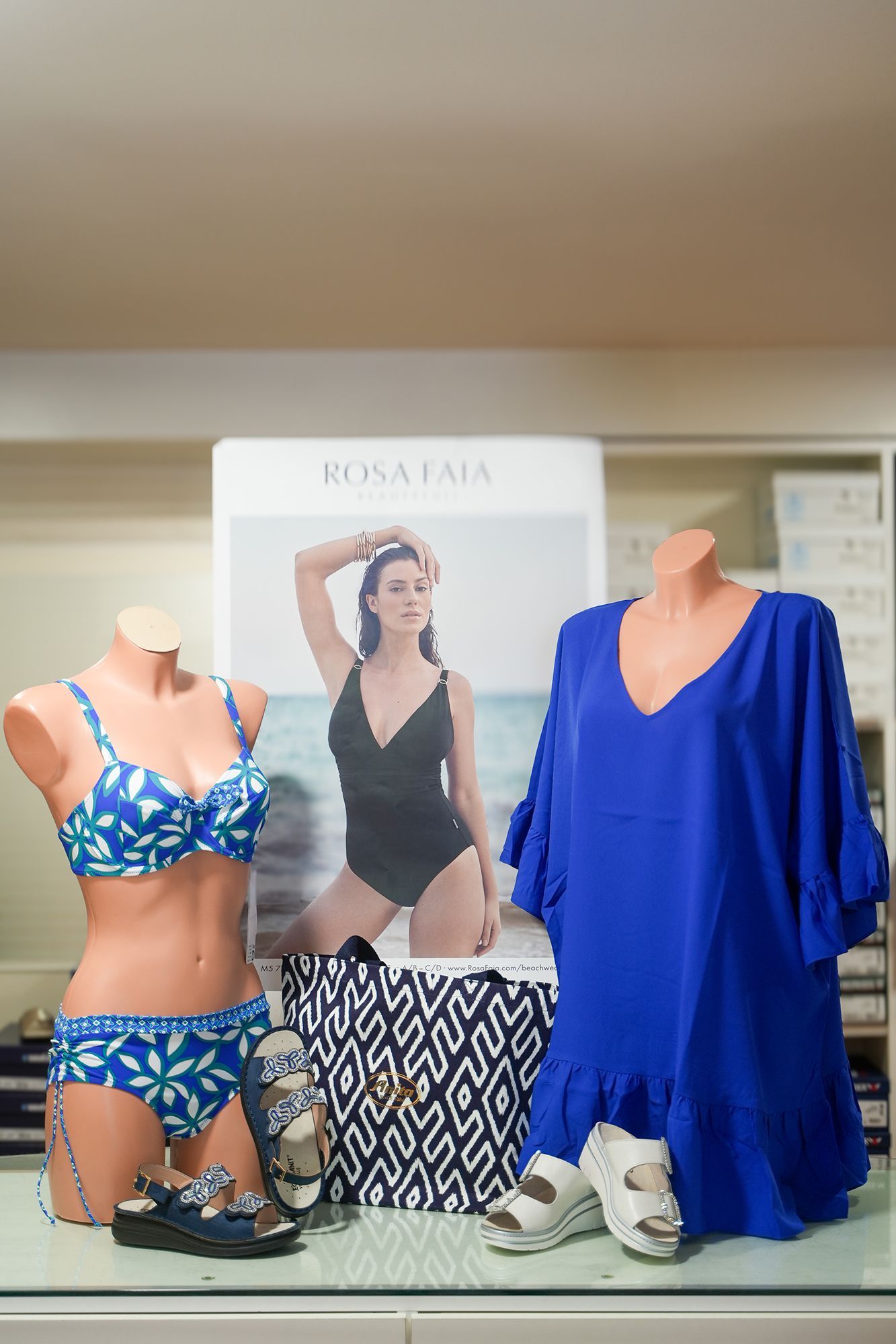 Costumi da bagno e copricostumi esposti su manichini con un poster in un negozio. Colori blu e bianco.