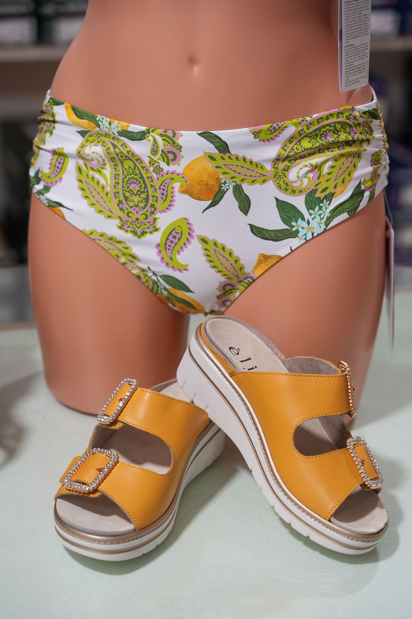 Manichino che indossa uno slip di bikini con fantasia bianca e verde, con sandali gialli con plateau.