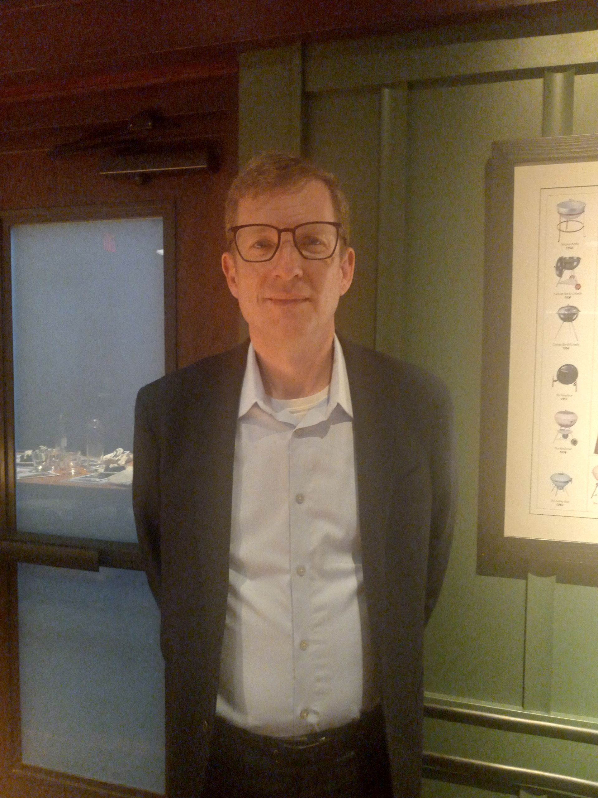 A man wearing glasses and a suit is standing in front of a picture on a wall.