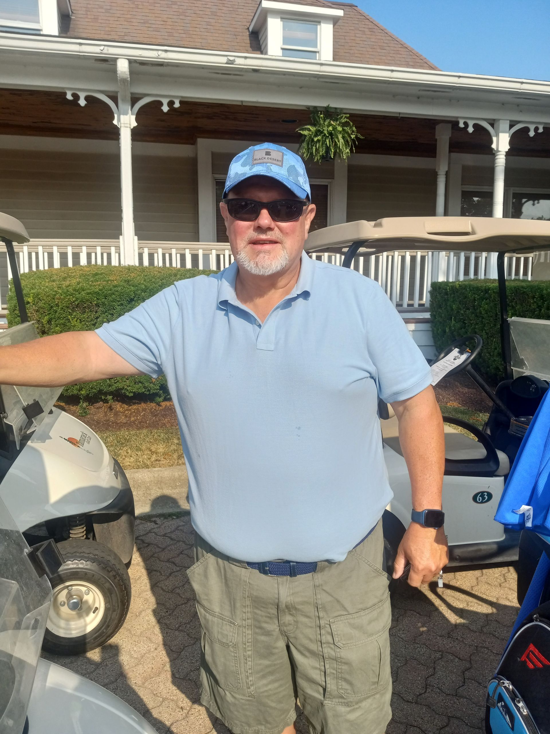 A man in a blue shirt and hat is standing in front of a golf cart.