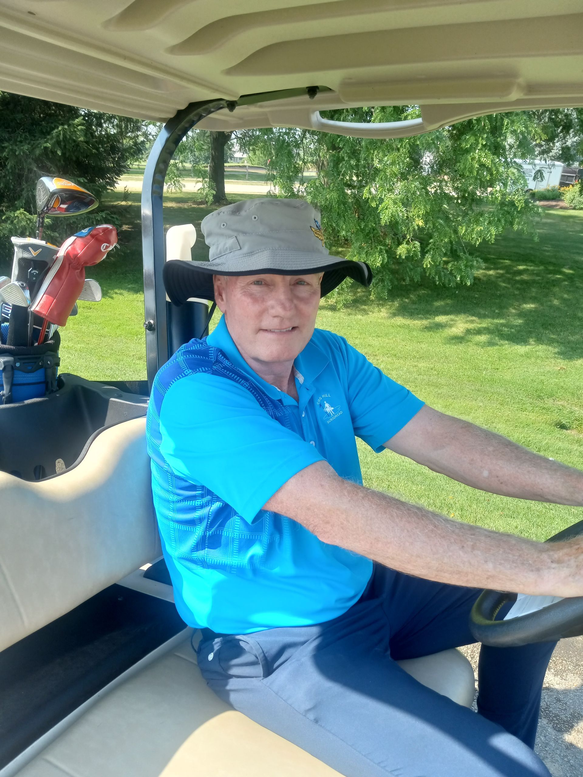 A man in a blue shirt and hat is driving a golf cart.