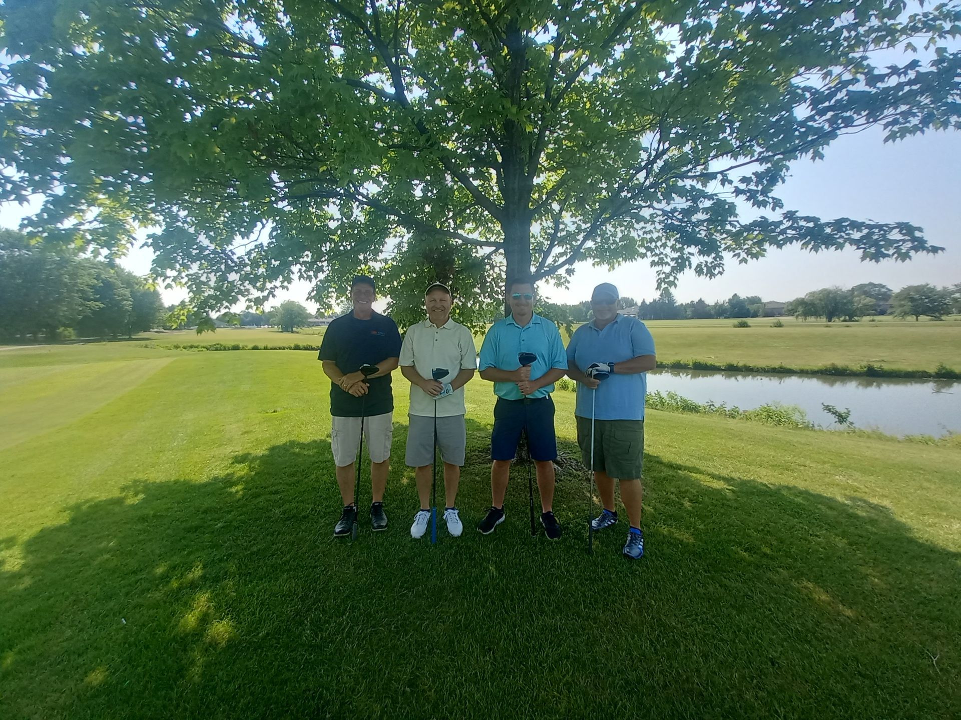 A group of men are standing under a tree on a golf course.