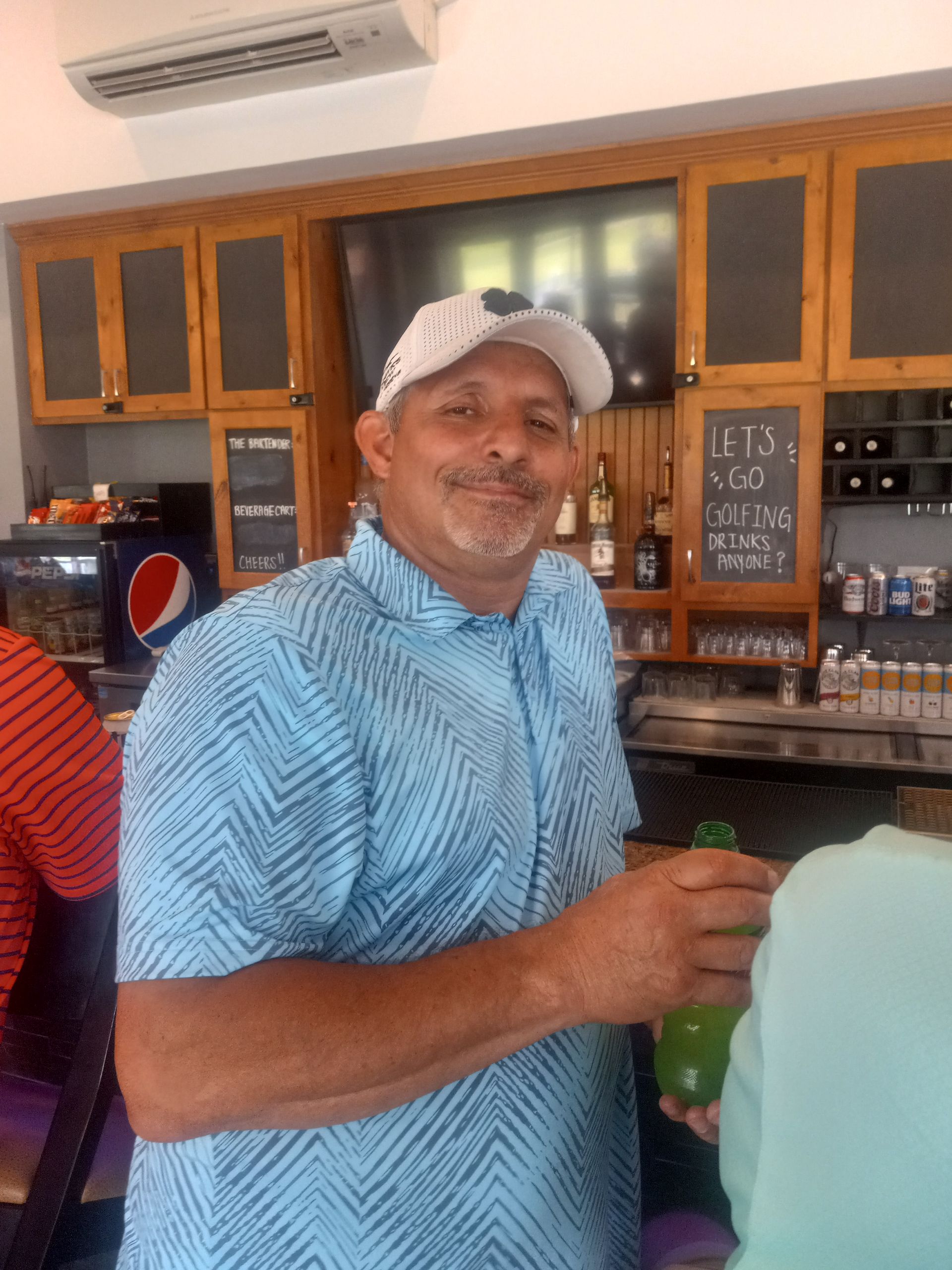 A man in a blue shirt and white hat is standing in a kitchen holding a drink.