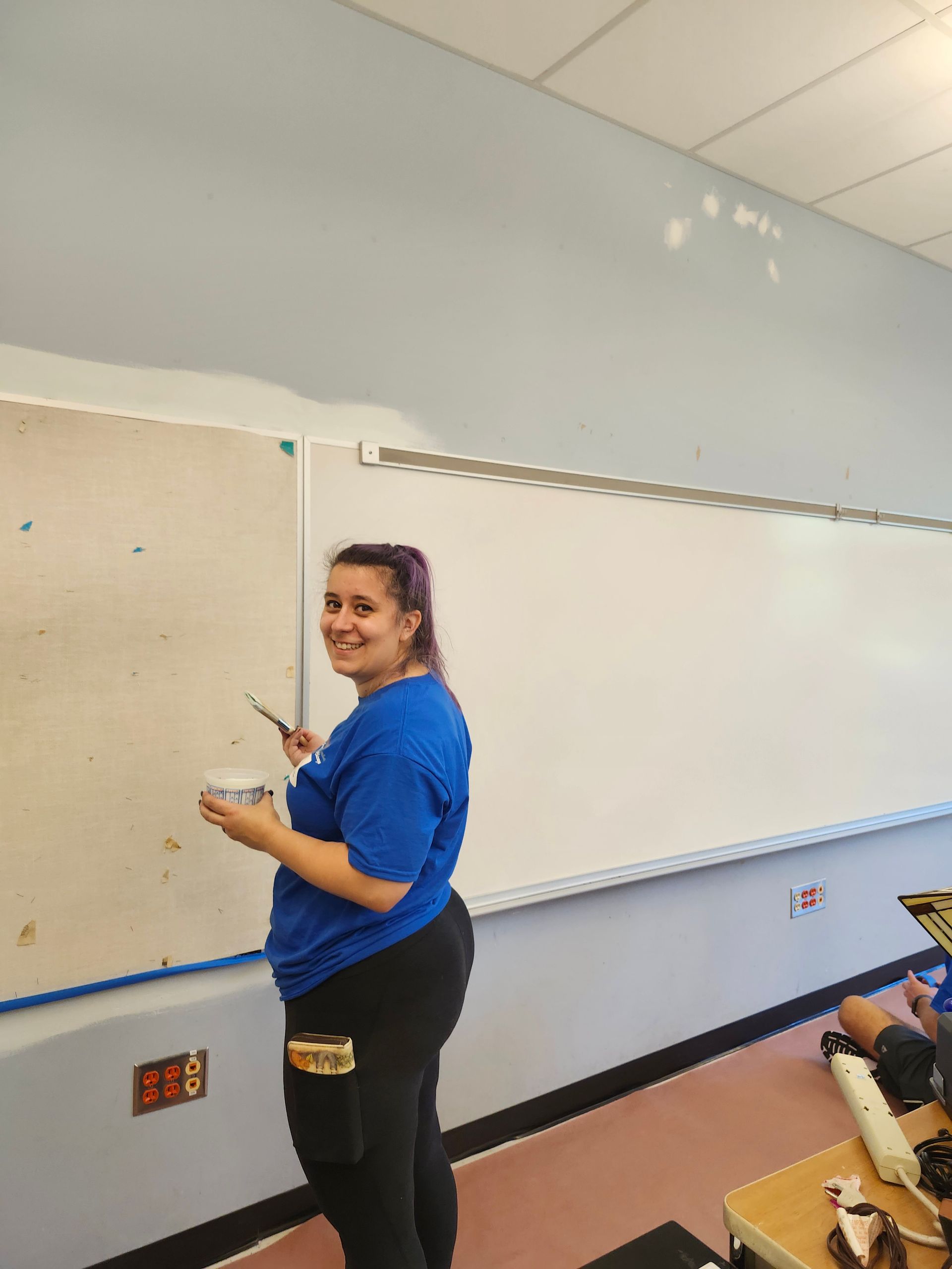 A woman in a blue shirt is painting a whiteboard in a classroom.