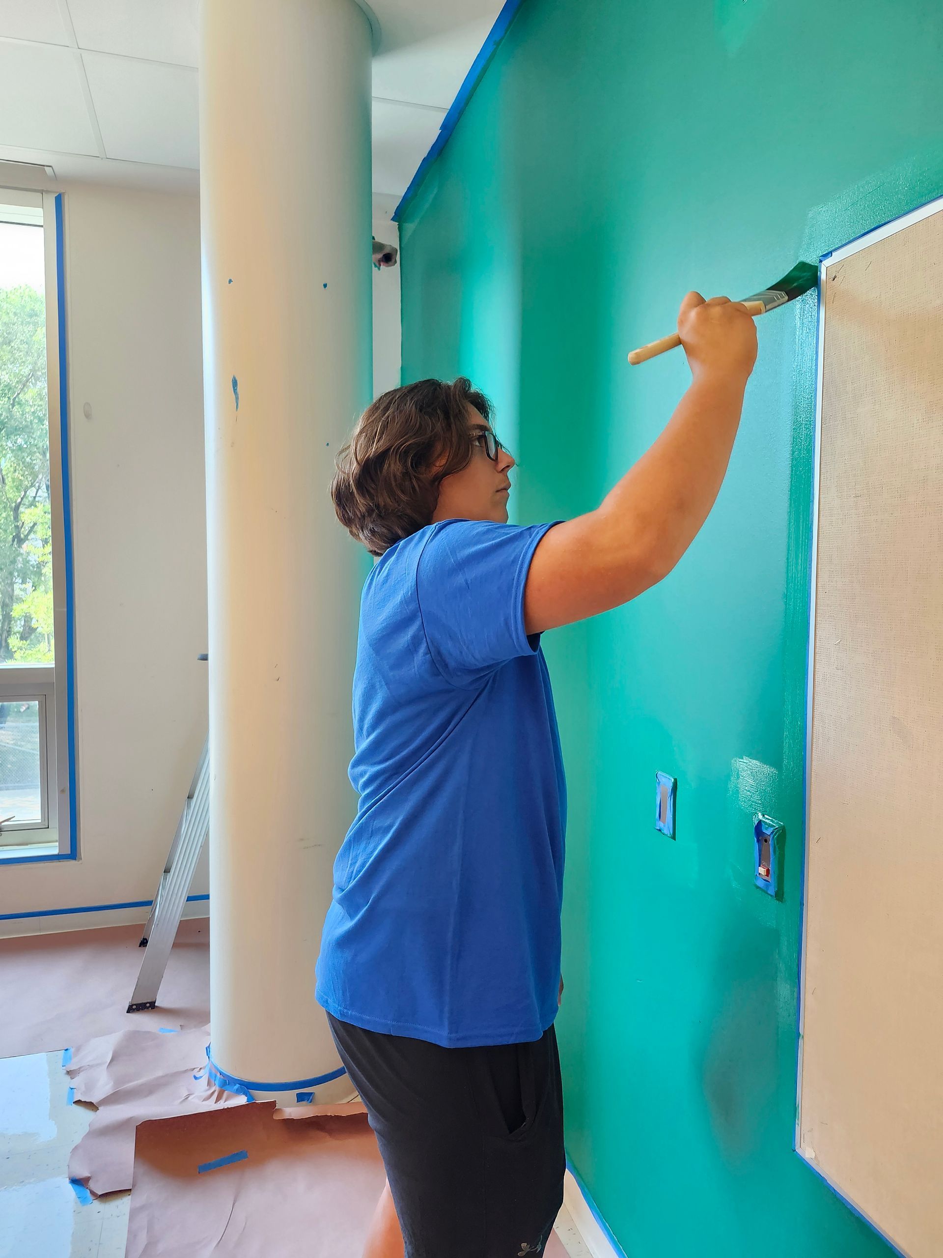 A woman in a blue shirt is painting a green wall with a brush.