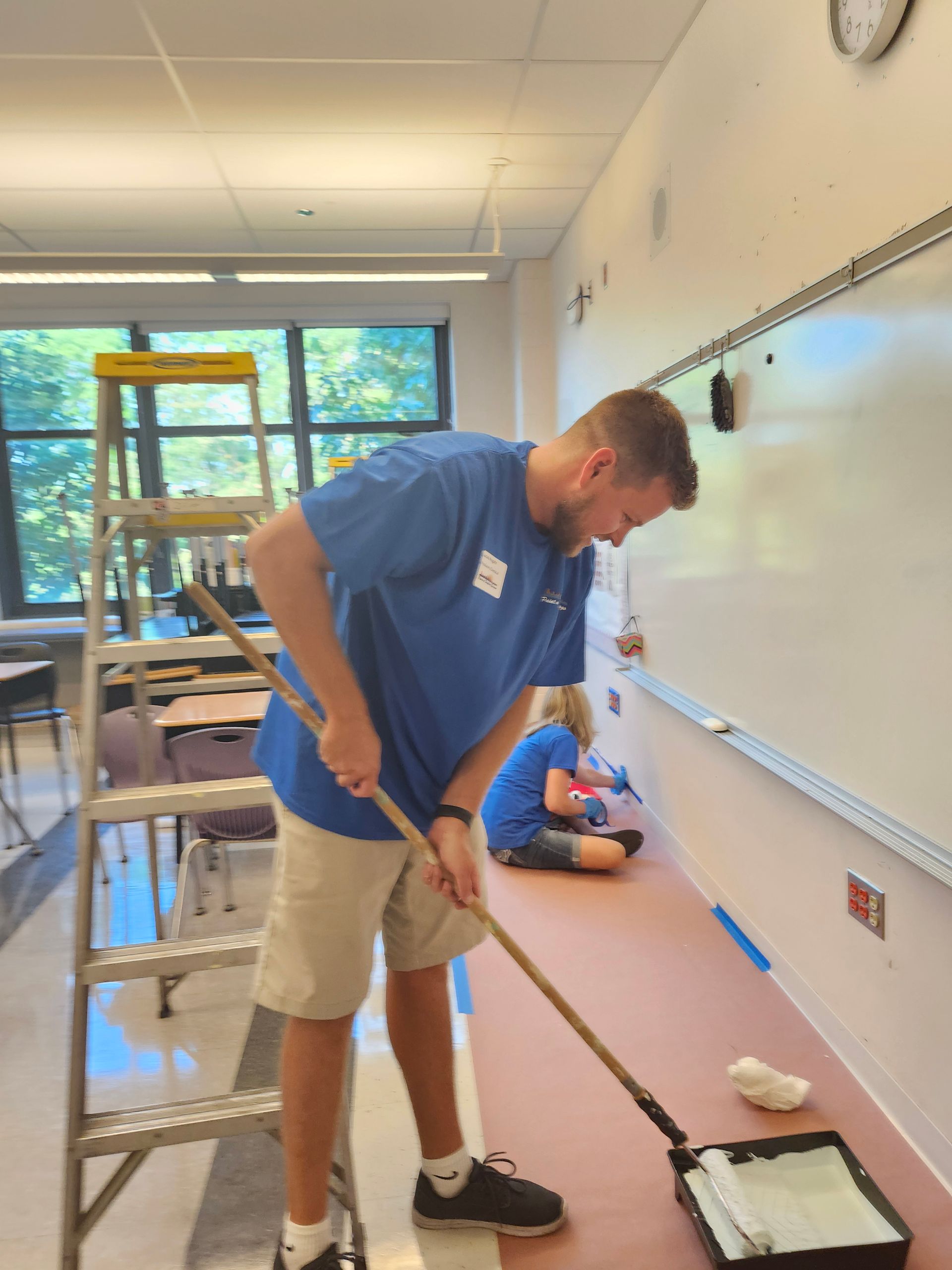 A man in a blue shirt is painting a wall in a classroom