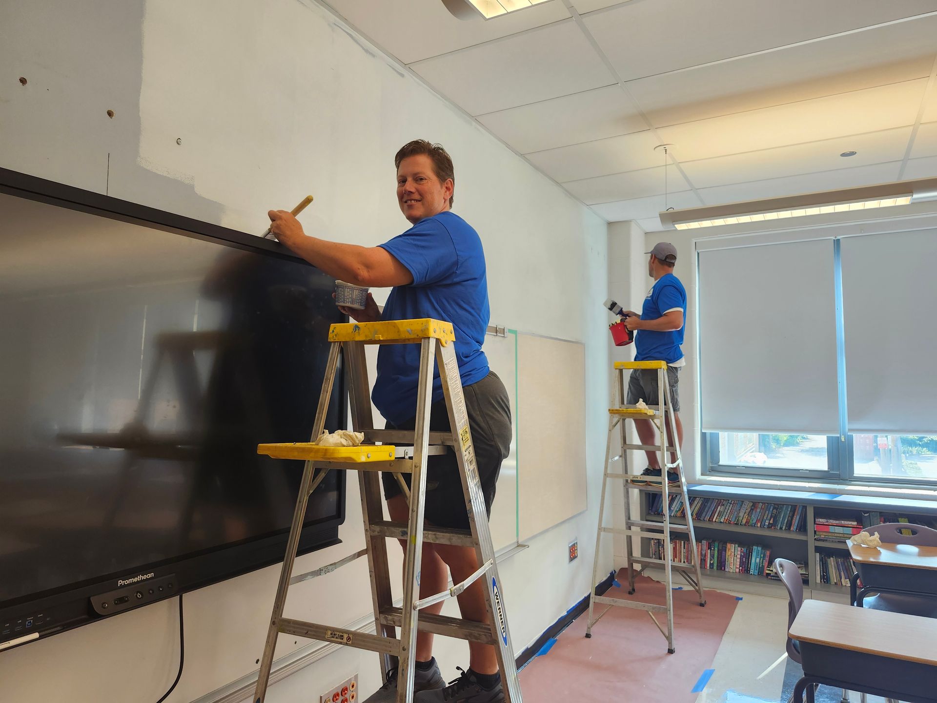 Two men are painting a blackboard in a classroom.