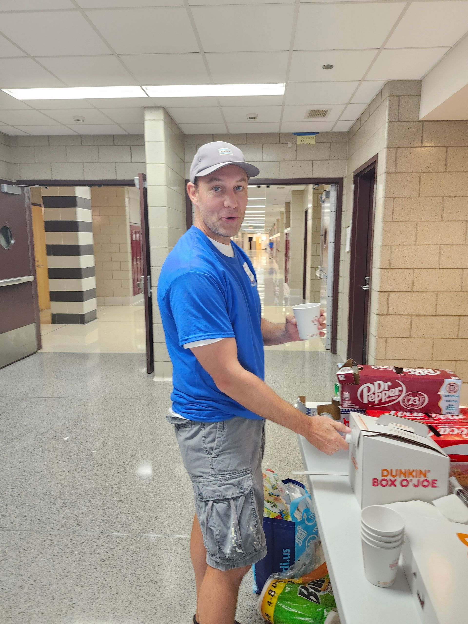 A man in a blue shirt is standing in a hallway holding a cup.