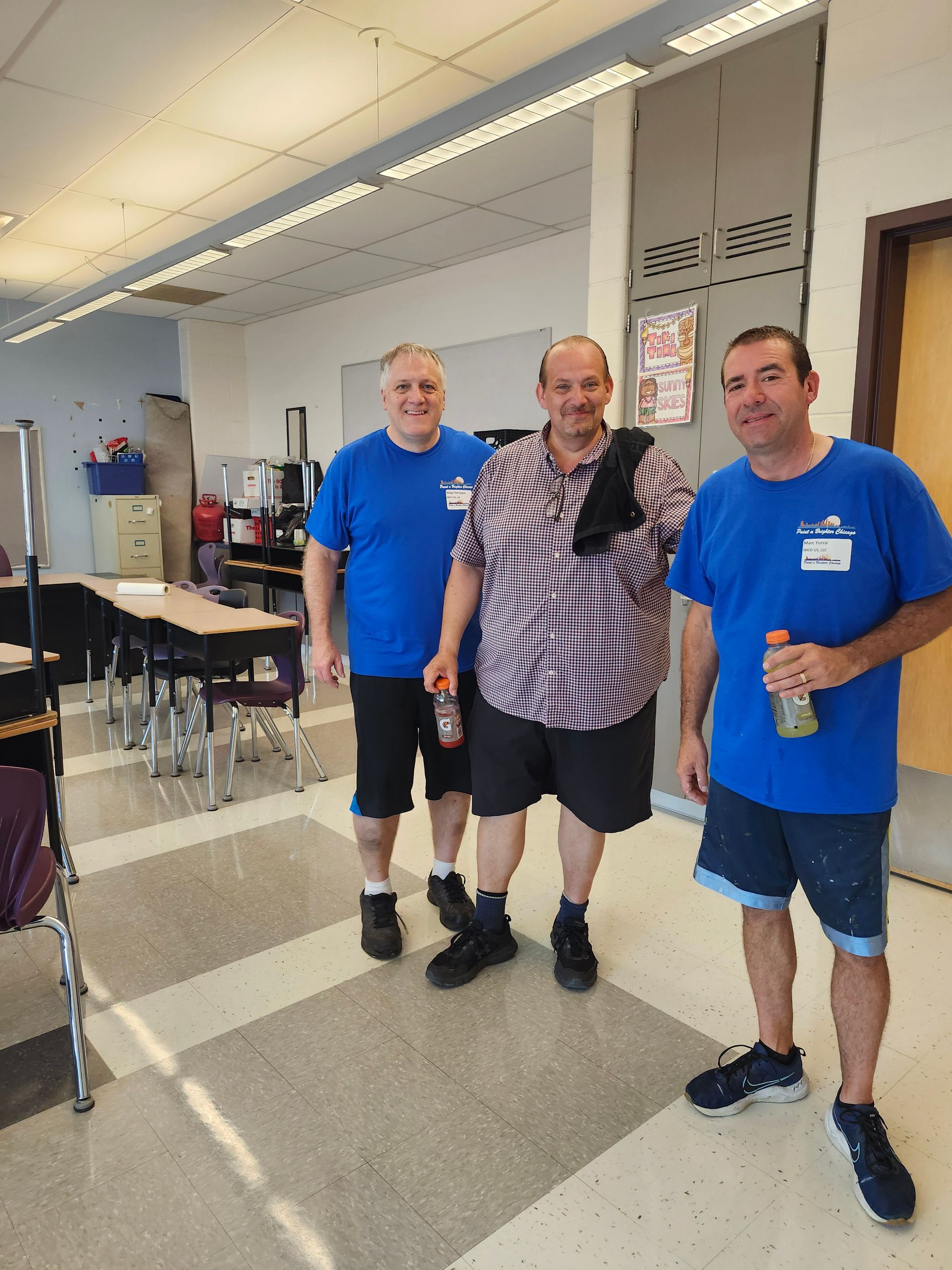 Three men in blue shirts are posing for a picture in a classroom.