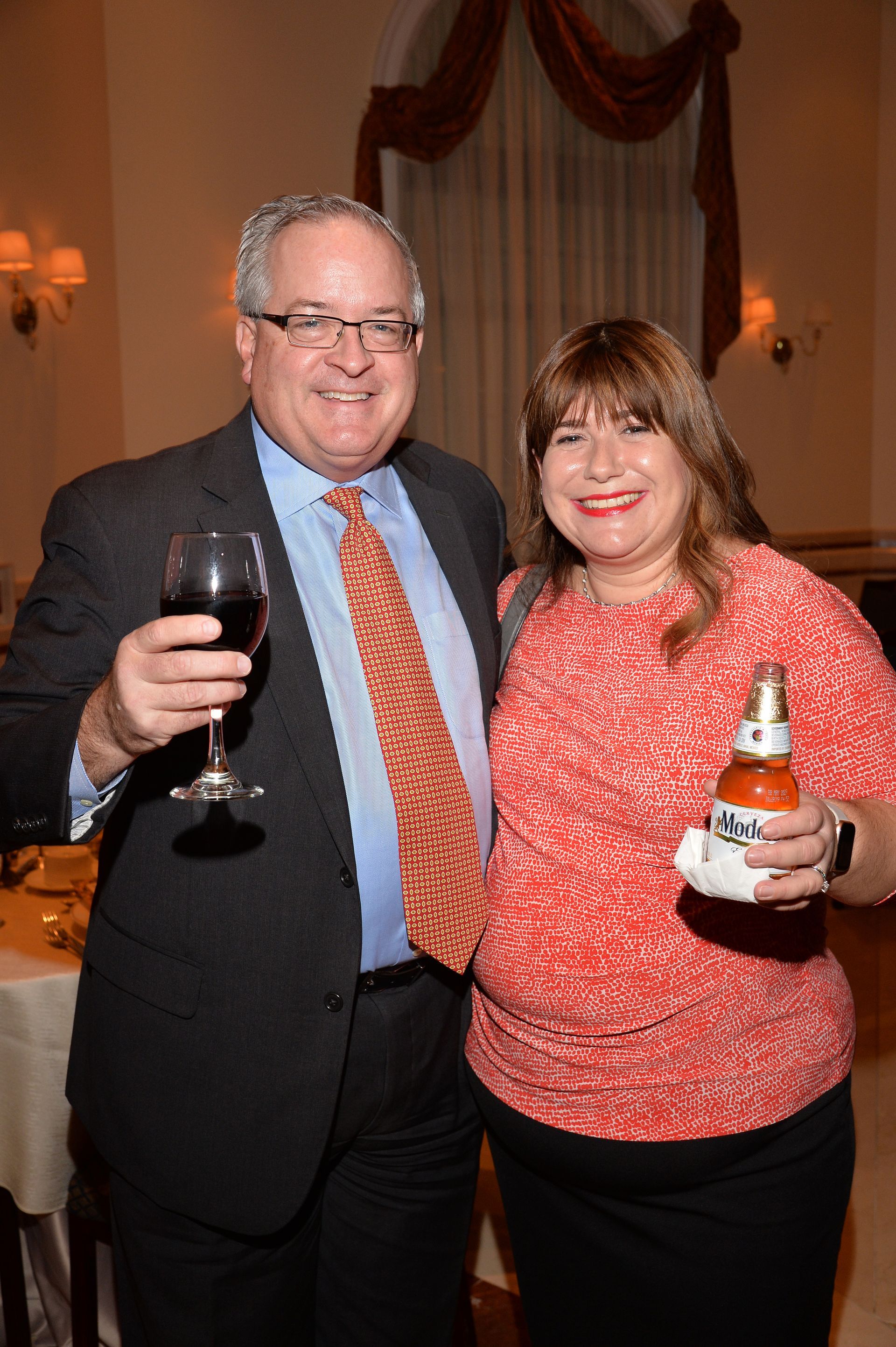 A man and a woman are posing for a picture while holding wine glasses and beer bottles.
