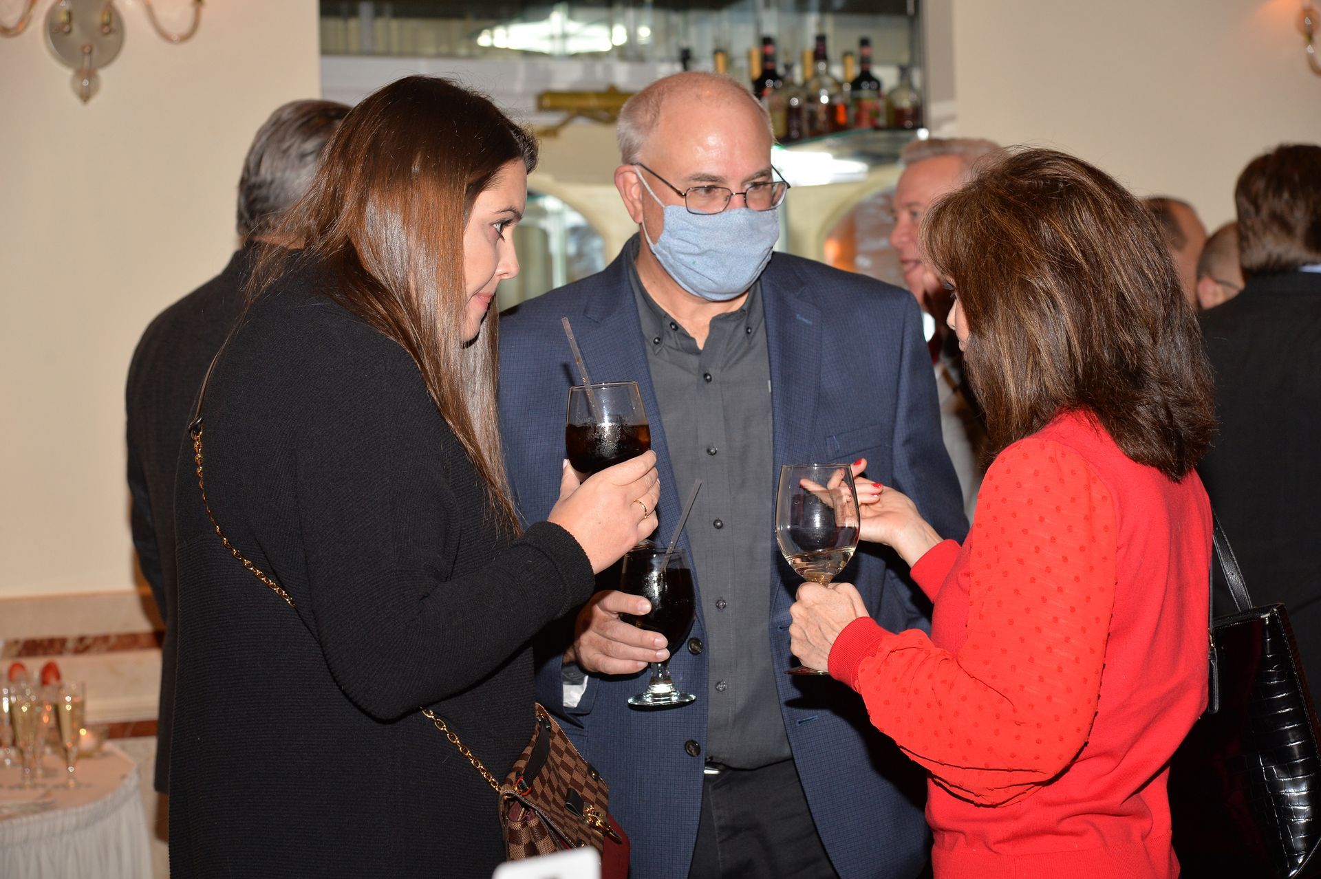 A man wearing a mask is talking to two women while holding wine glasses.