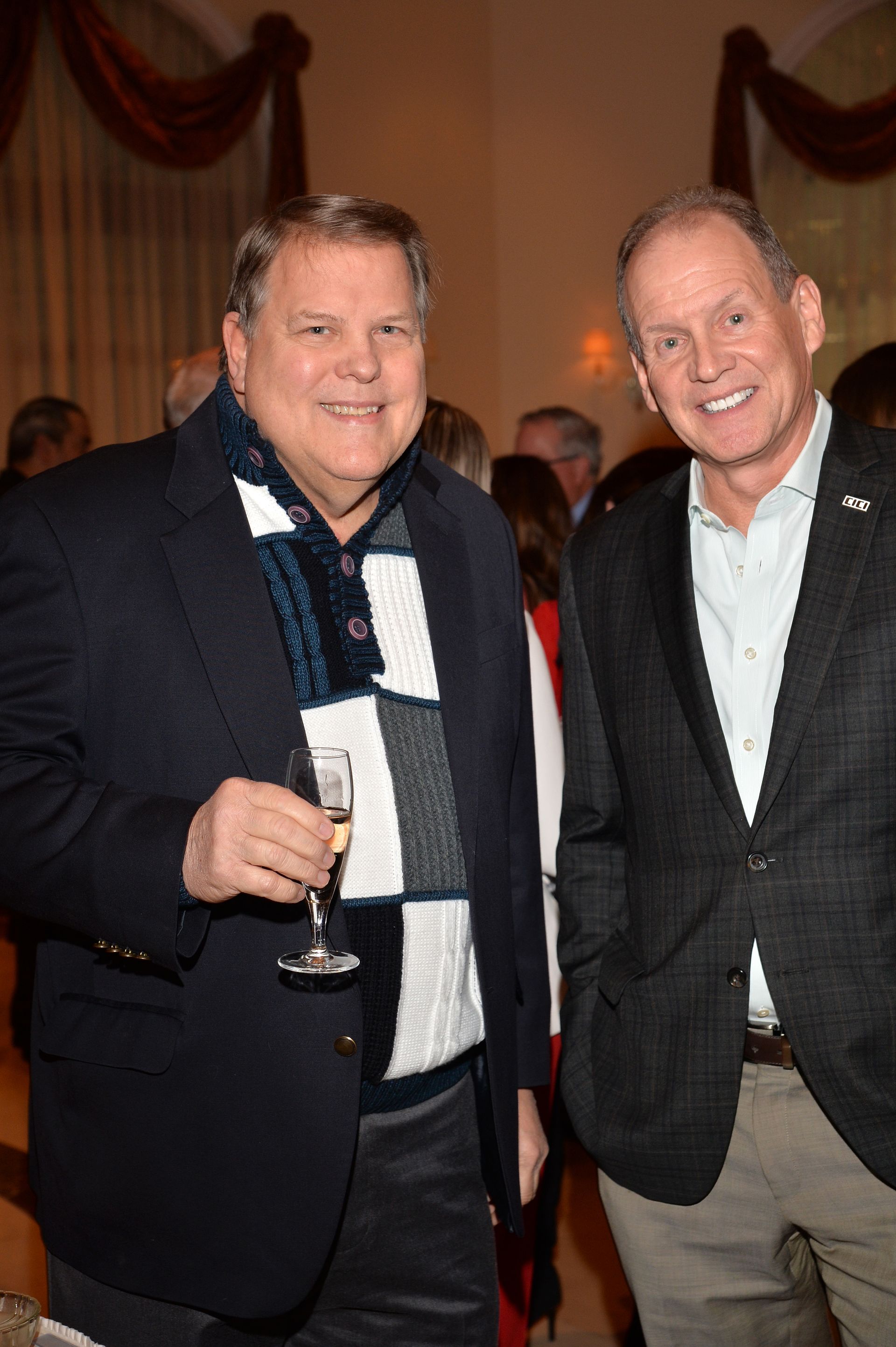 Two men are standing next to each other in a room and one is holding a glass of wine.