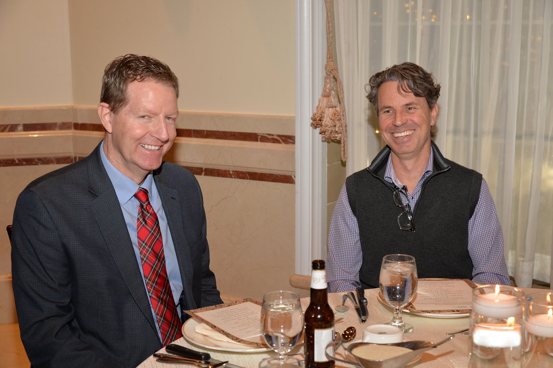 Two men are sitting at a table with wine glasses and a bottle of beer.