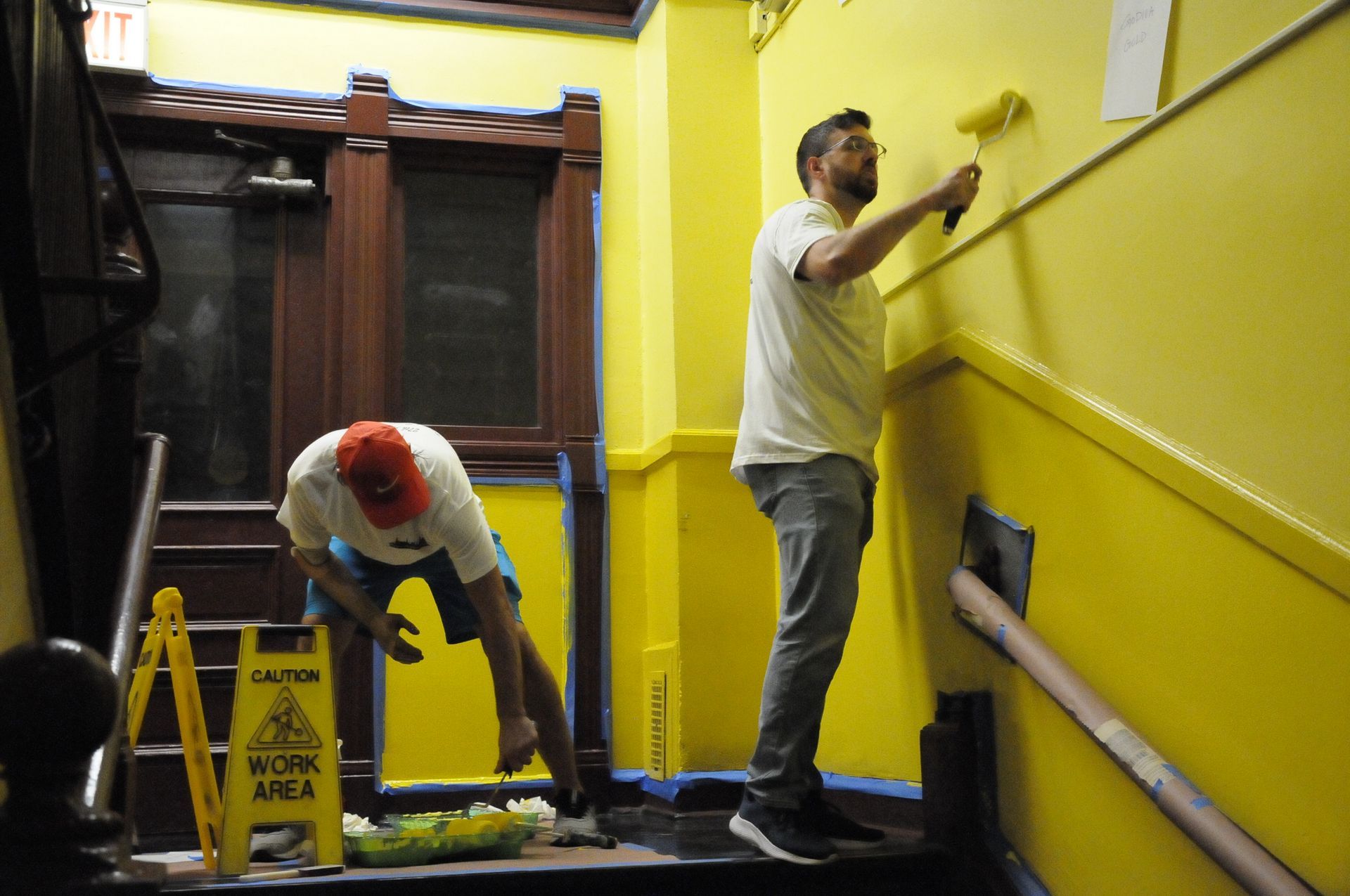 Two men are painting a yellow wall next to a caution sign