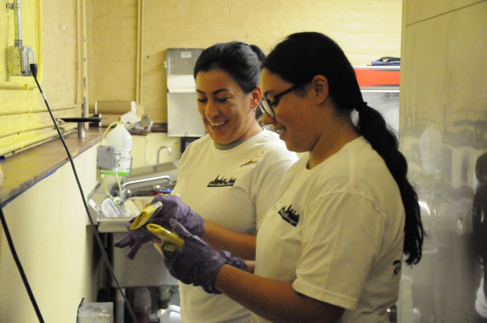 Two women wearing white shirts and purple gloves are working in a kitchen