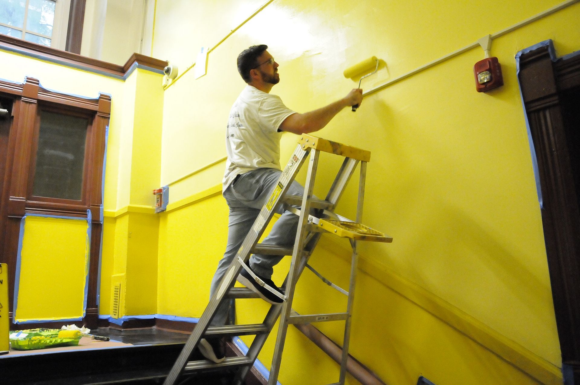 A man is standing on a ladder painting a yellow wall.
