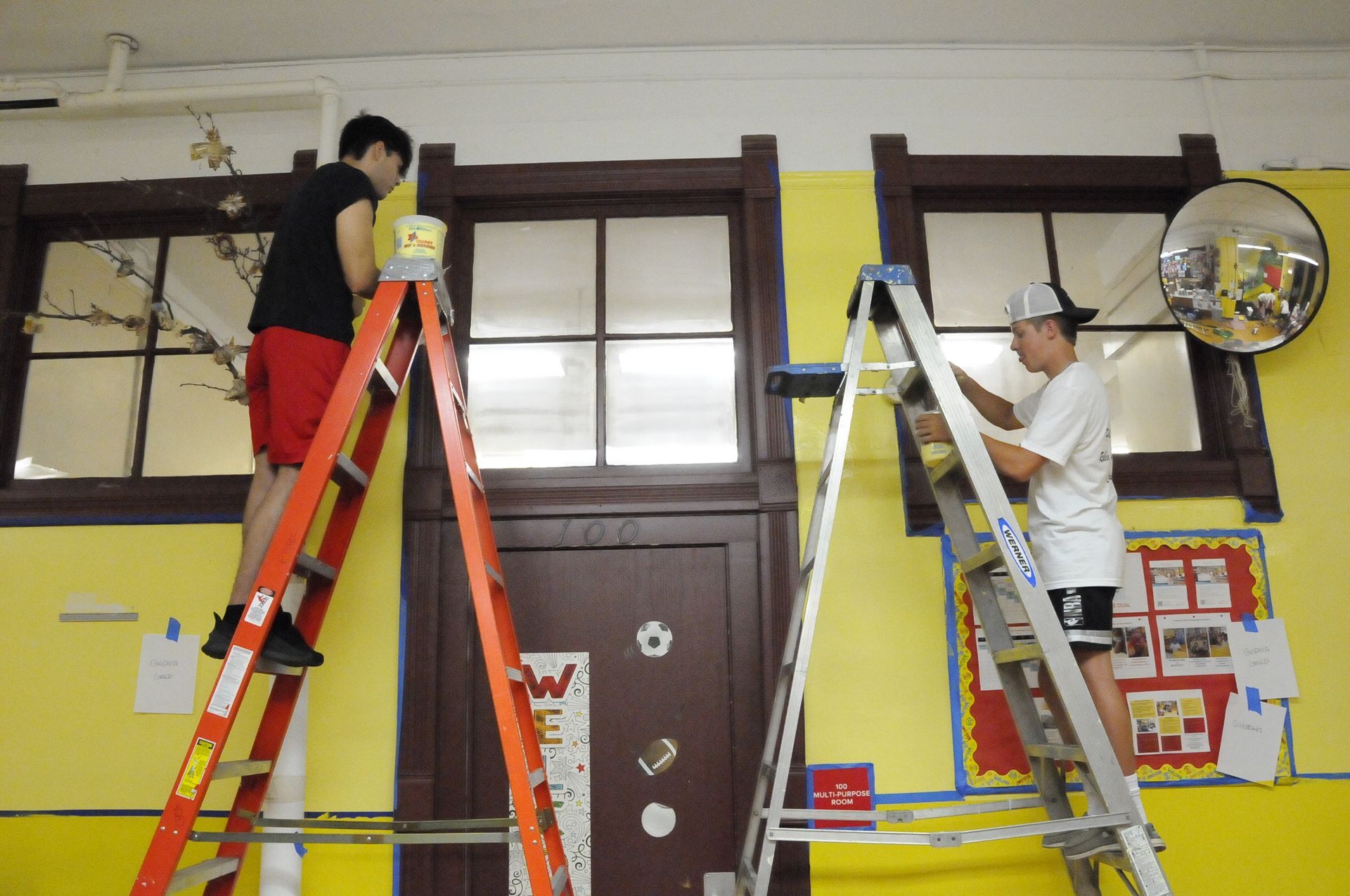 Two men on ladders paint a yellow wall