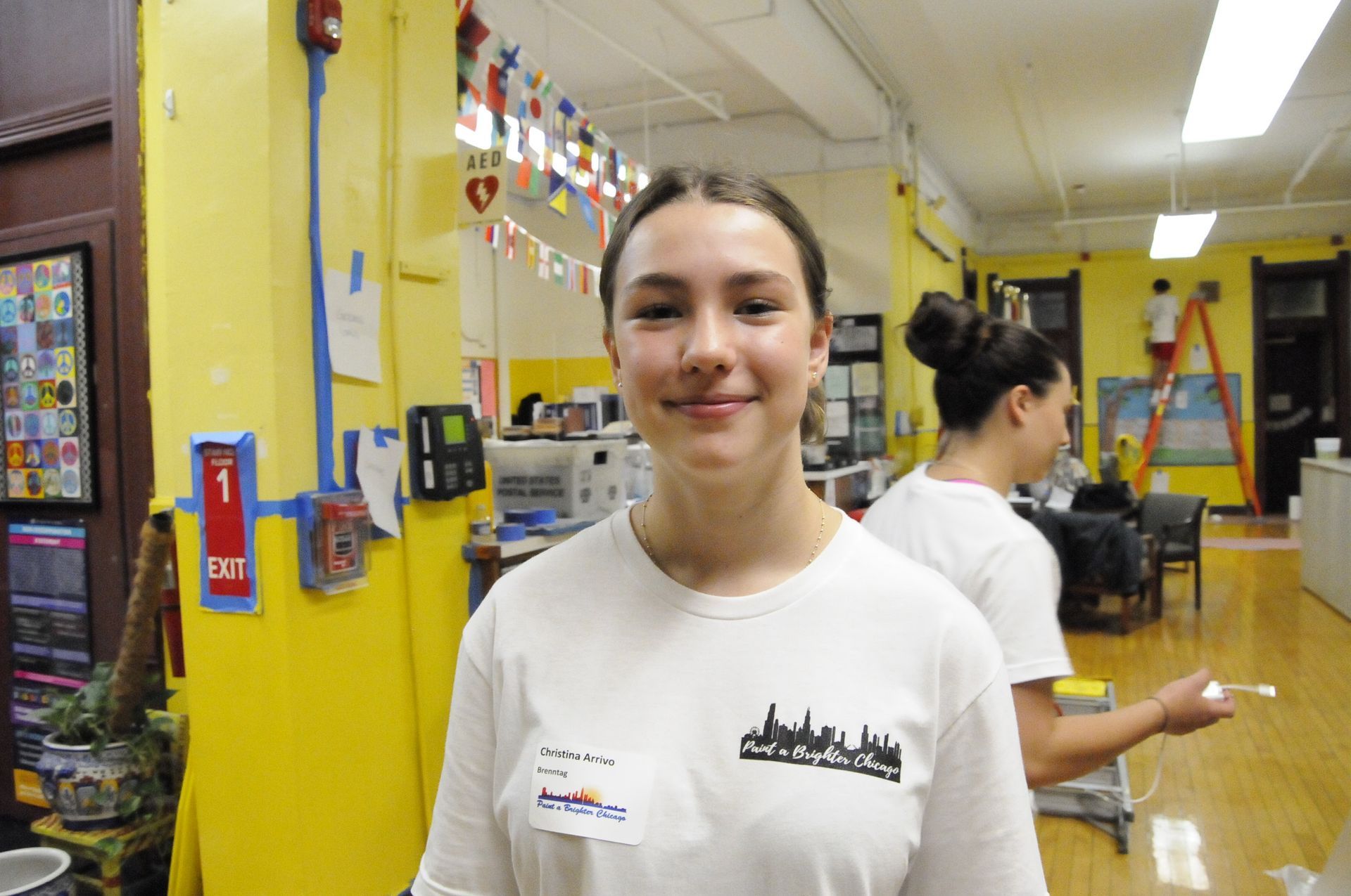 A woman in a white shirt is standing in front of a yellow wall.