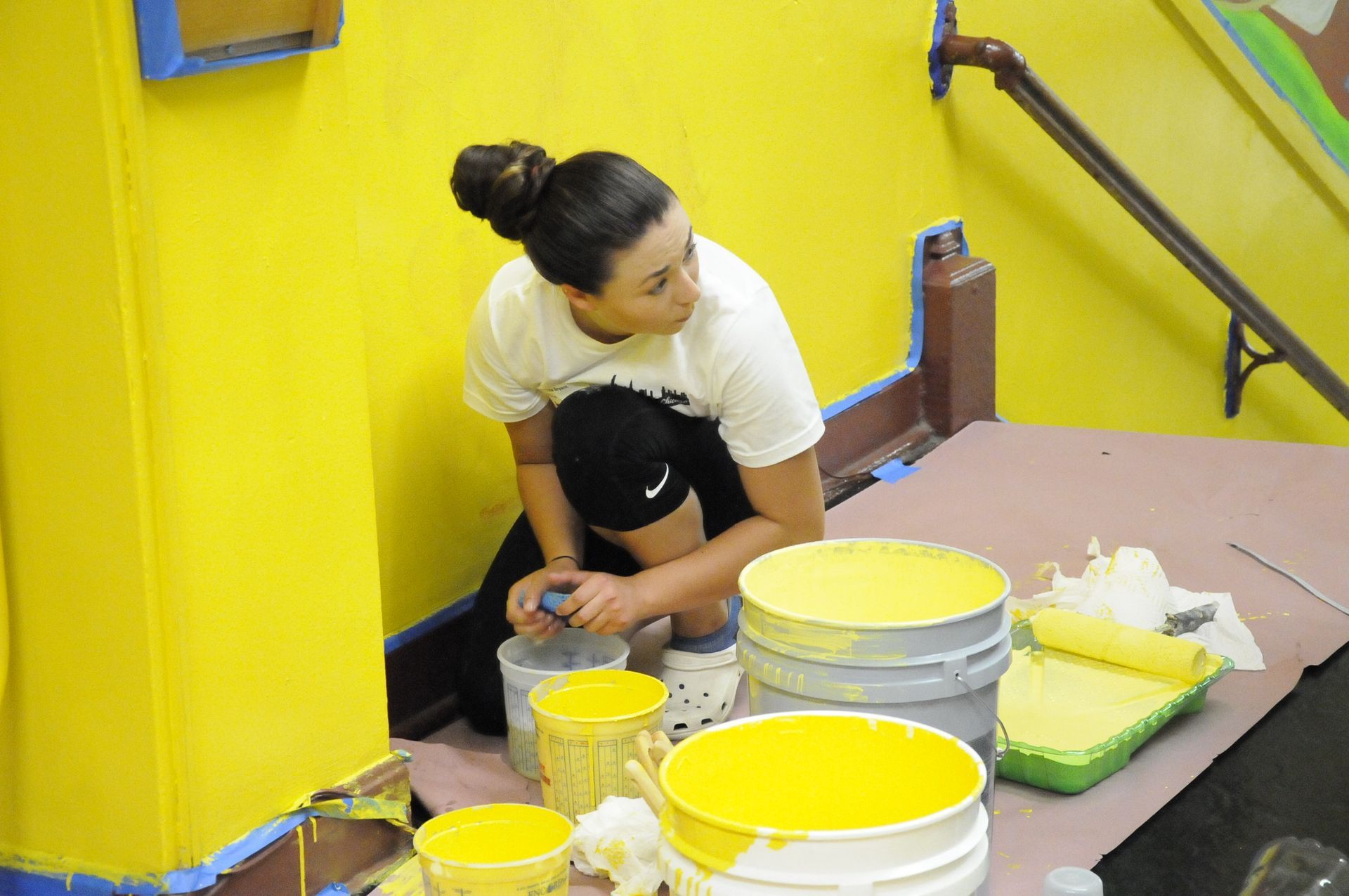 A woman is kneeling on the floor painting a yellow wall.