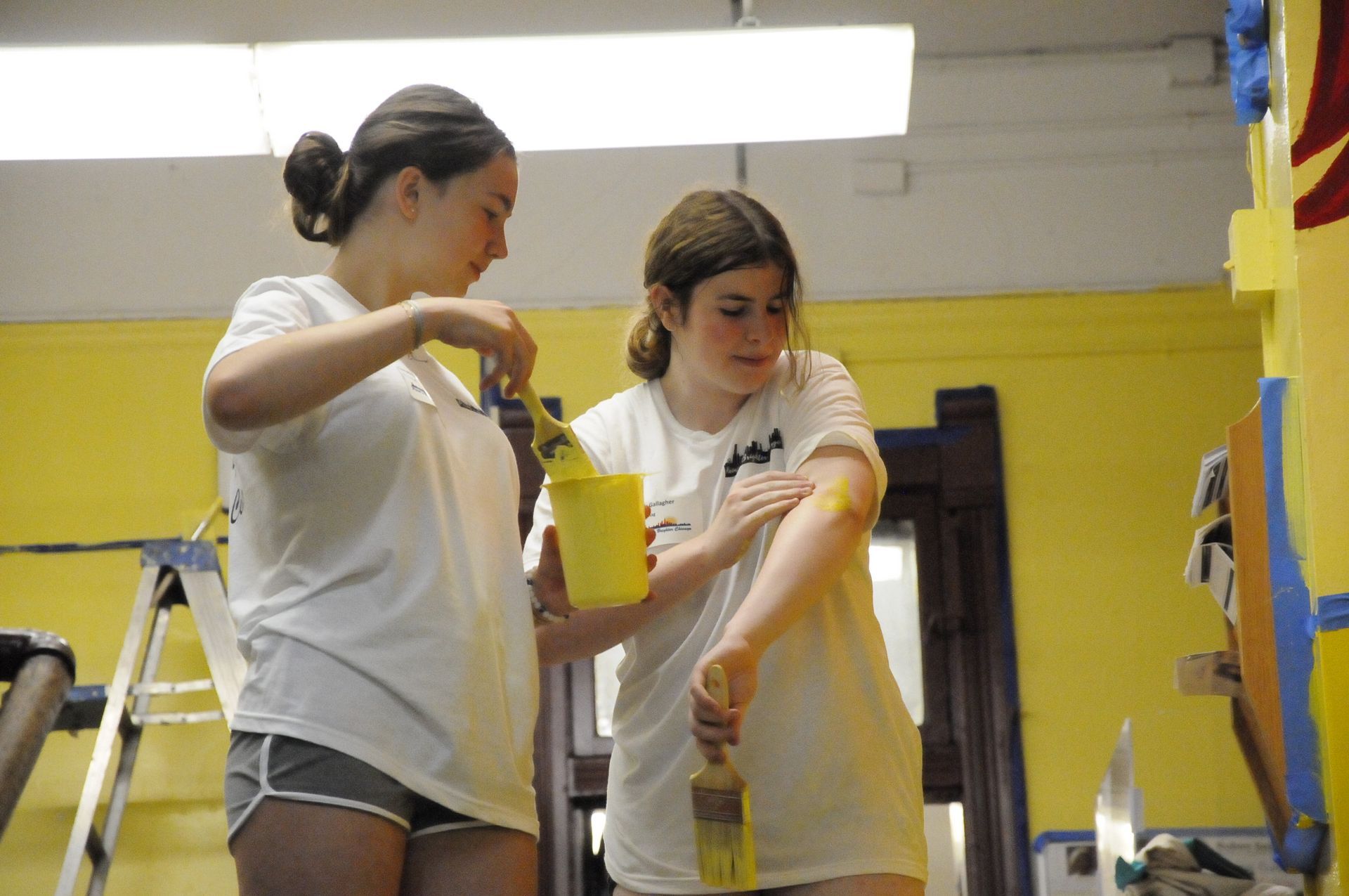 Two young women are painting a yellow wall together.