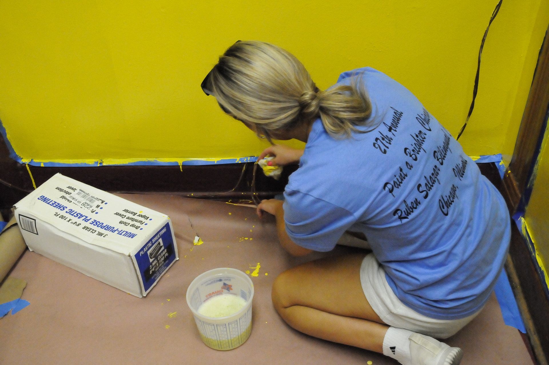 A woman in a blue shirt is kneeling down and painting a wall