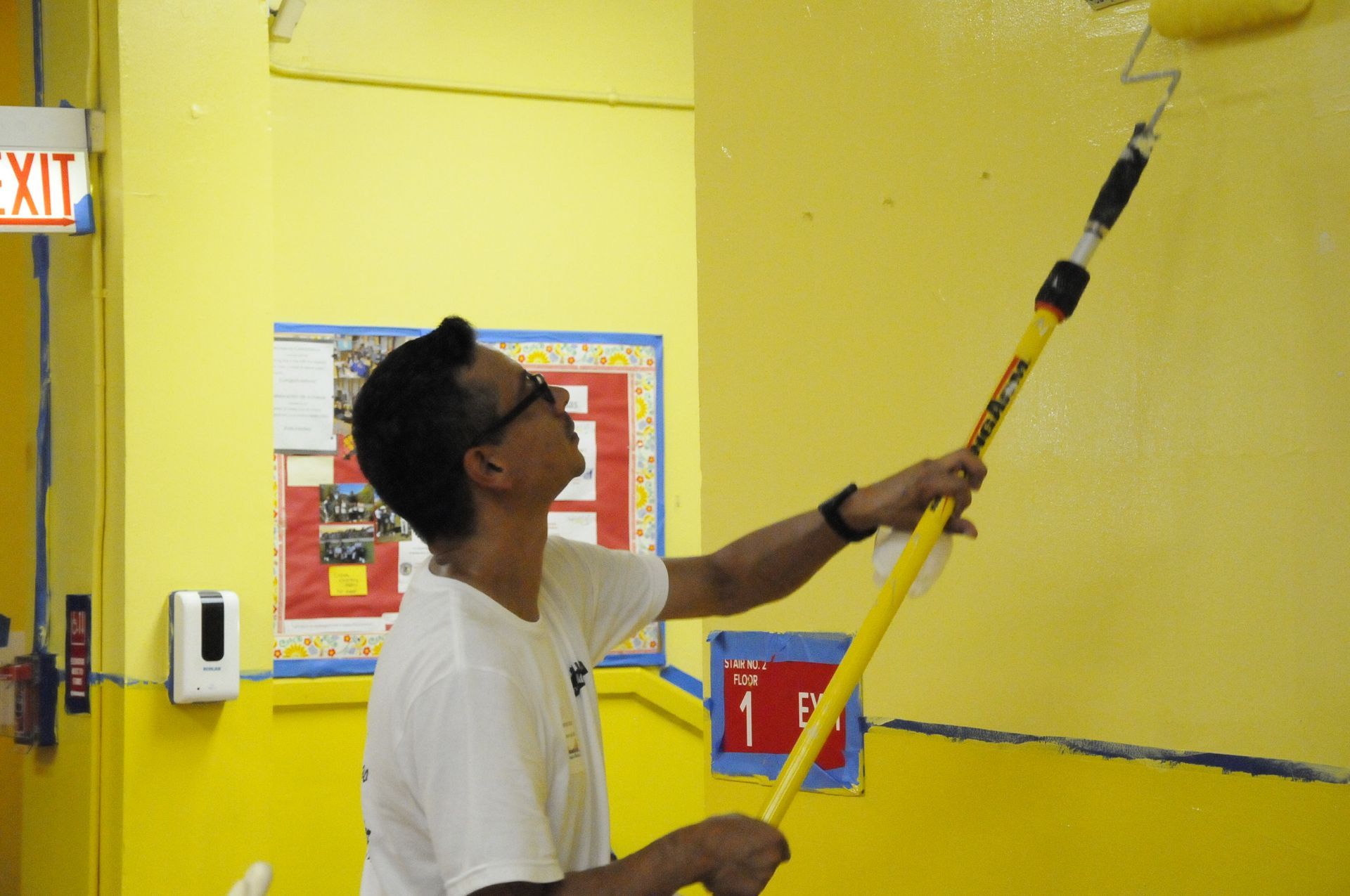 A man is painting a yellow wall in front of an exit sign