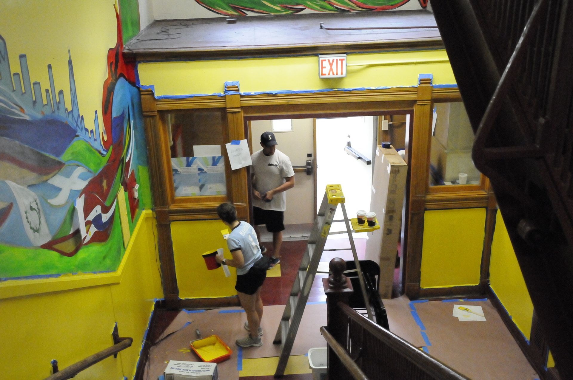 A man and a woman are painting a wall in front of an exit sign