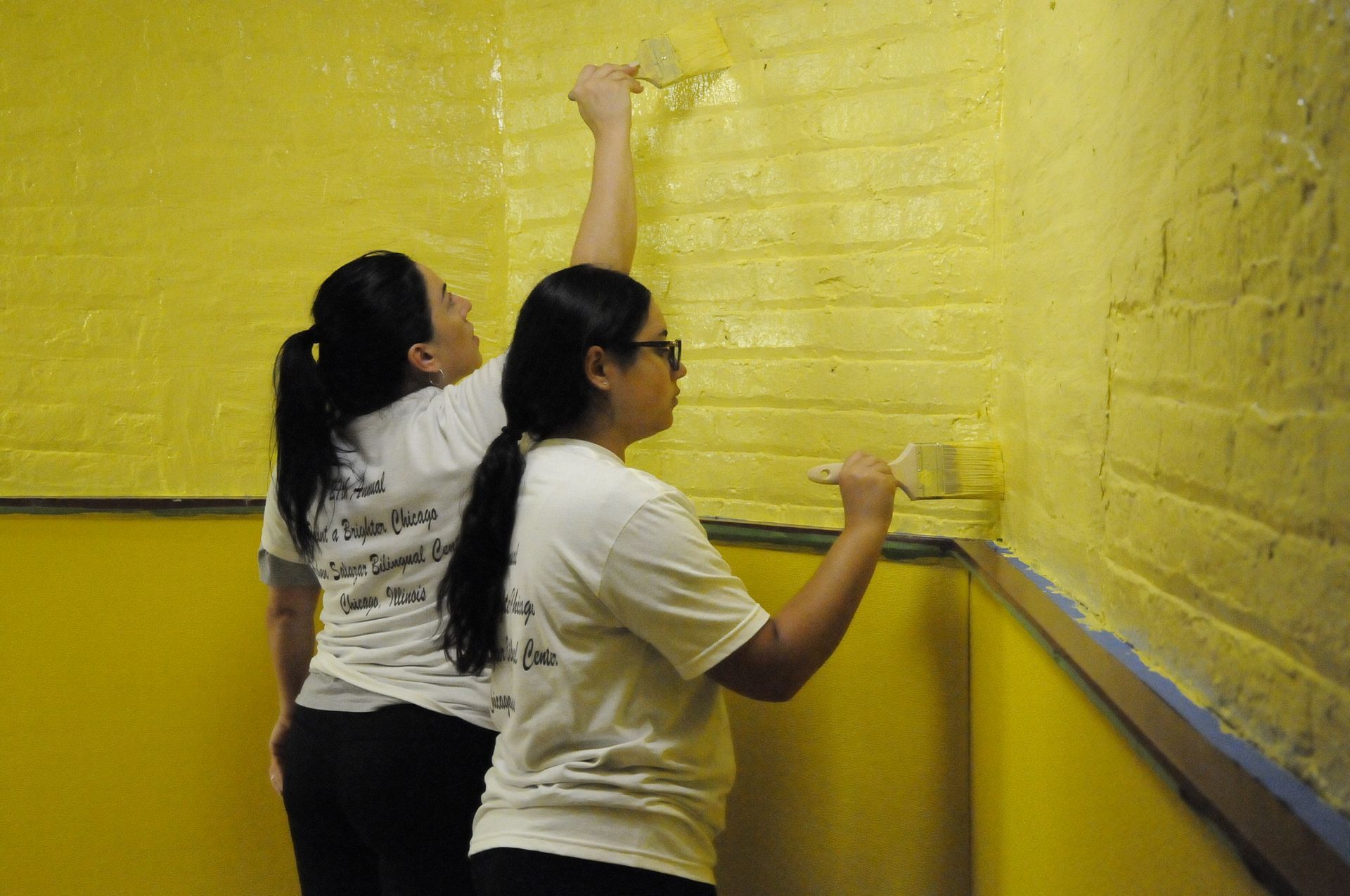 Two women are painting a yellow brick wall.