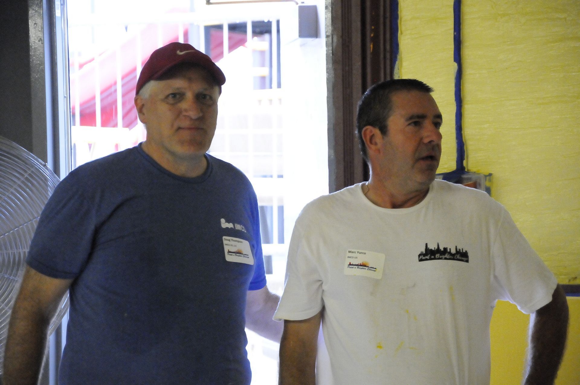 Two men are standing next to each other in front of a yellow wall