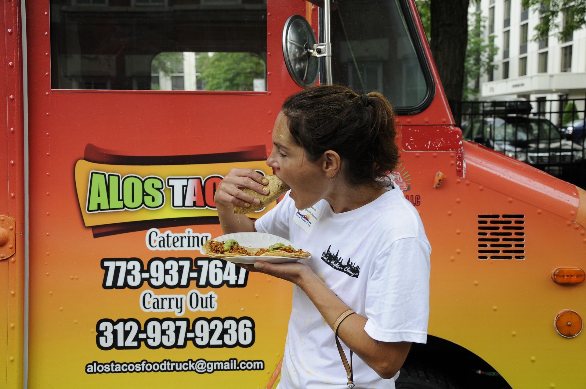 A woman is eating a sandwich in front of an alos taco truck