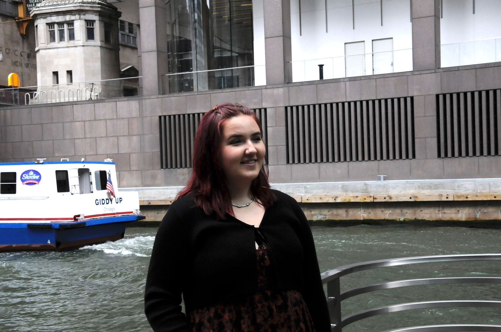 A woman stands in front of a river with a boat in the background