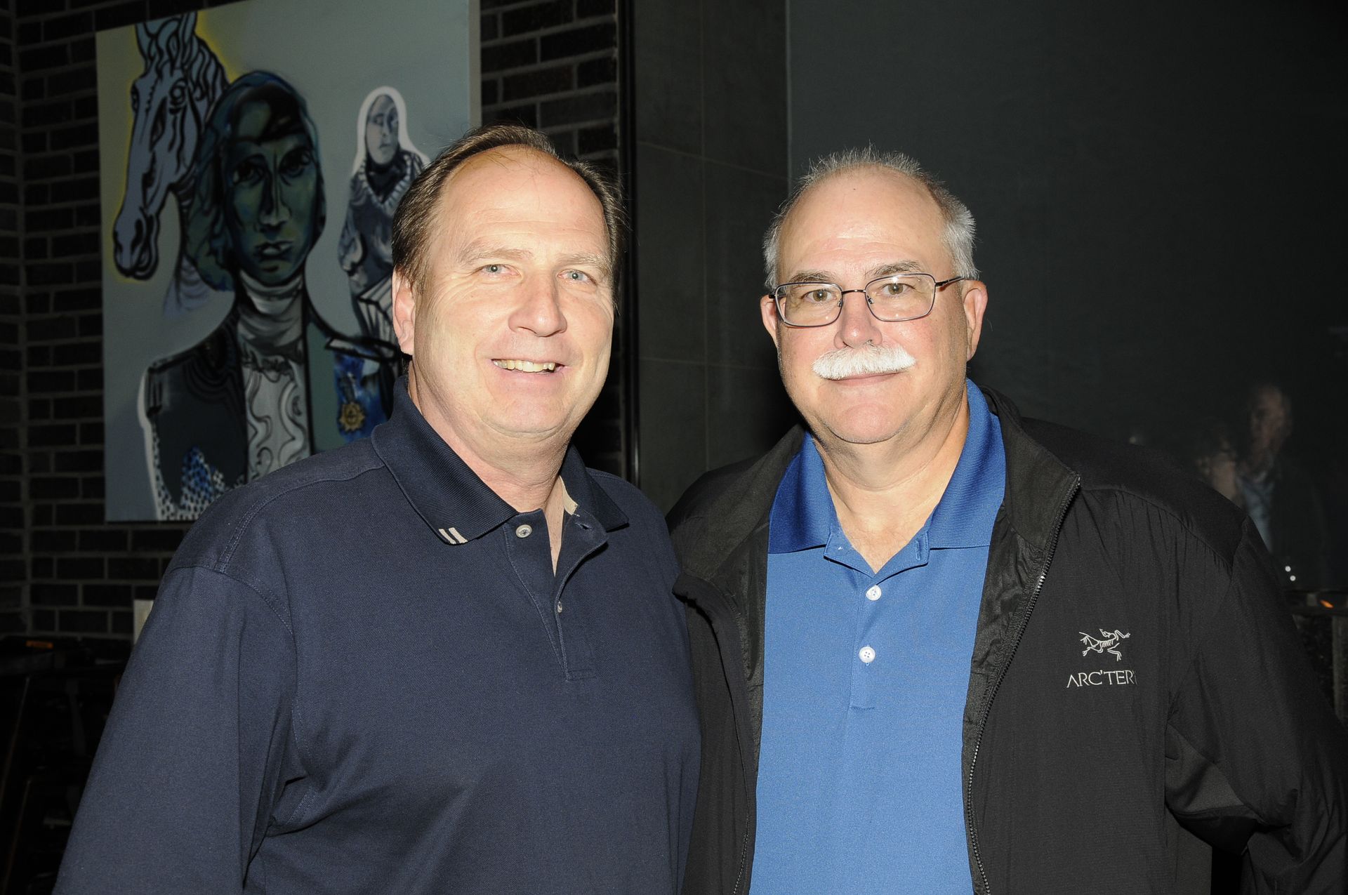 Two men are posing for a picture in front of a painting.