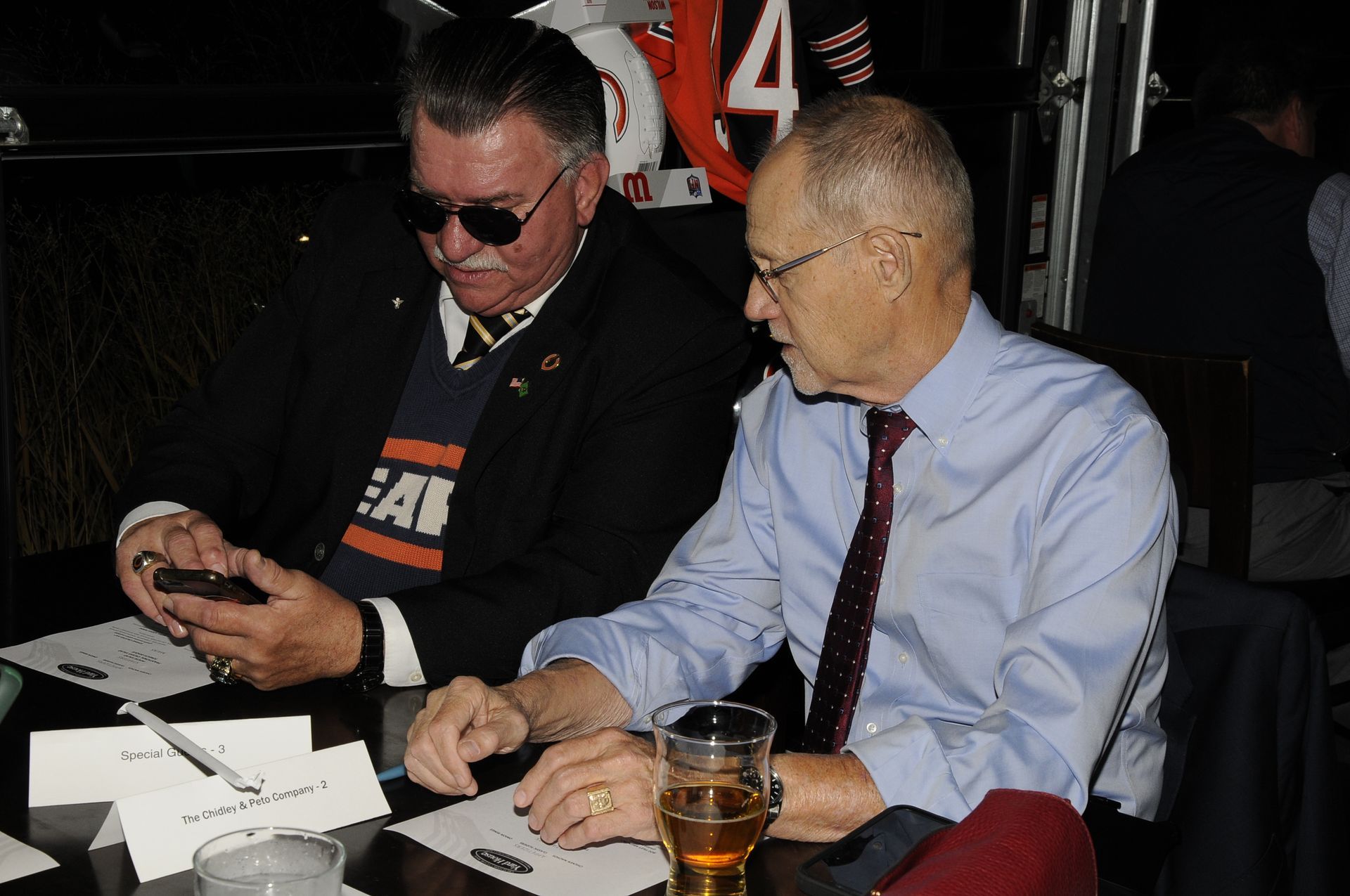 Two men sit at a table with one wearing a shirt that says eat