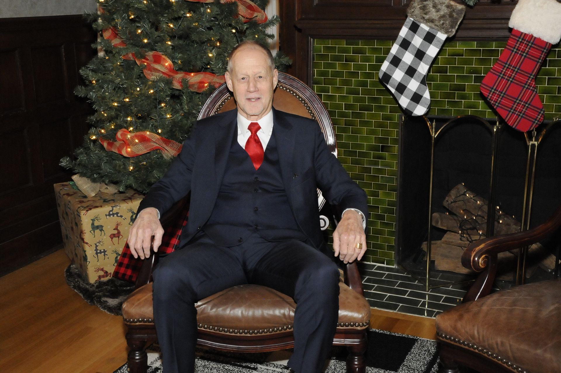 A man in a suit and tie is sitting in a chair in front of a christmas tree.