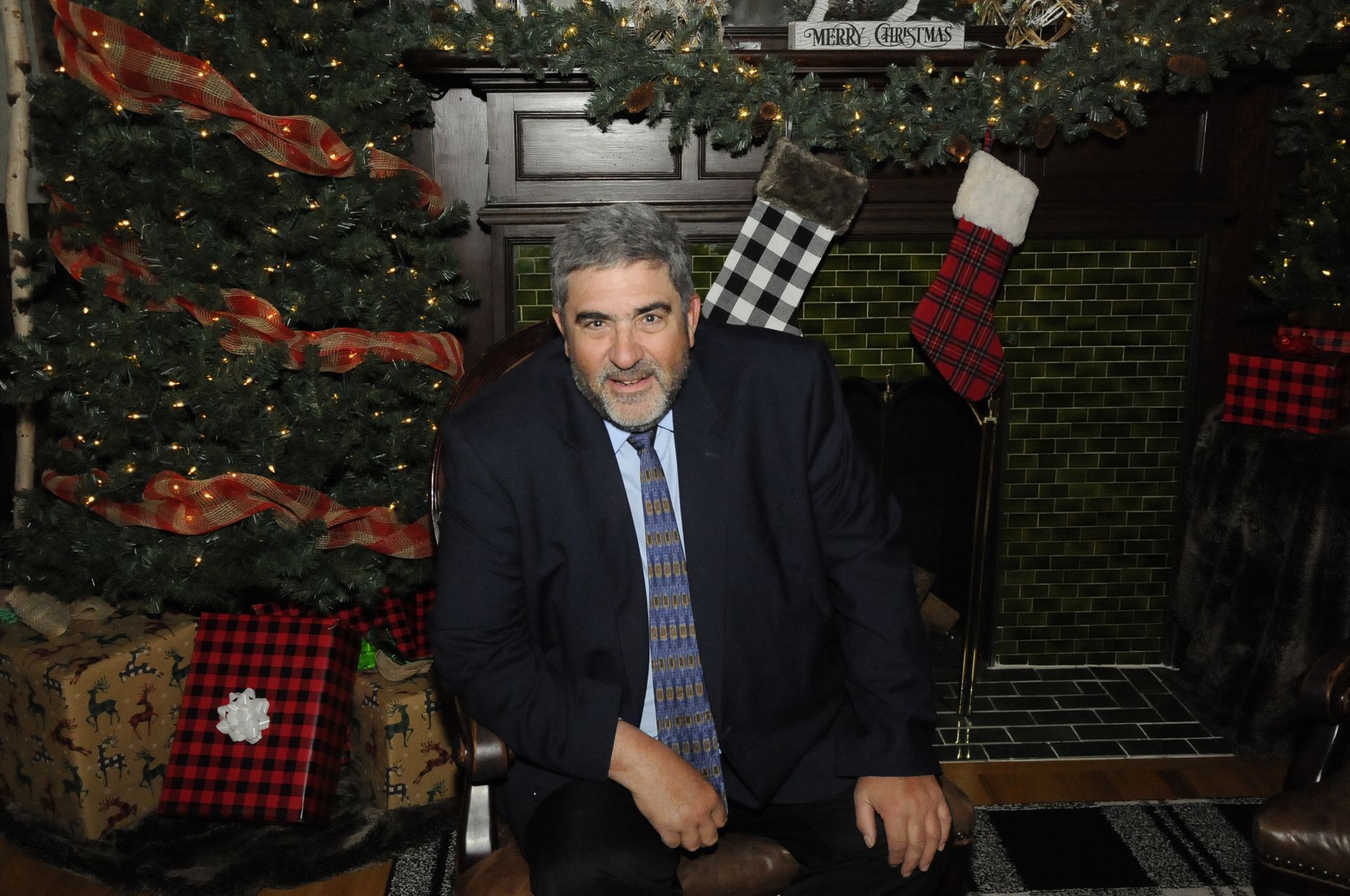 A man in a suit and tie is sitting in front of a christmas tree.