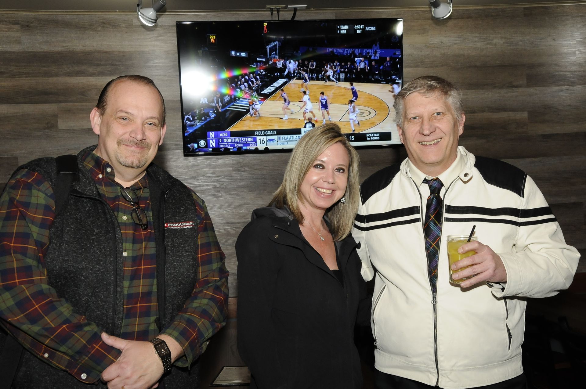 Three people are posing for a picture in front of a basketball game on a television.