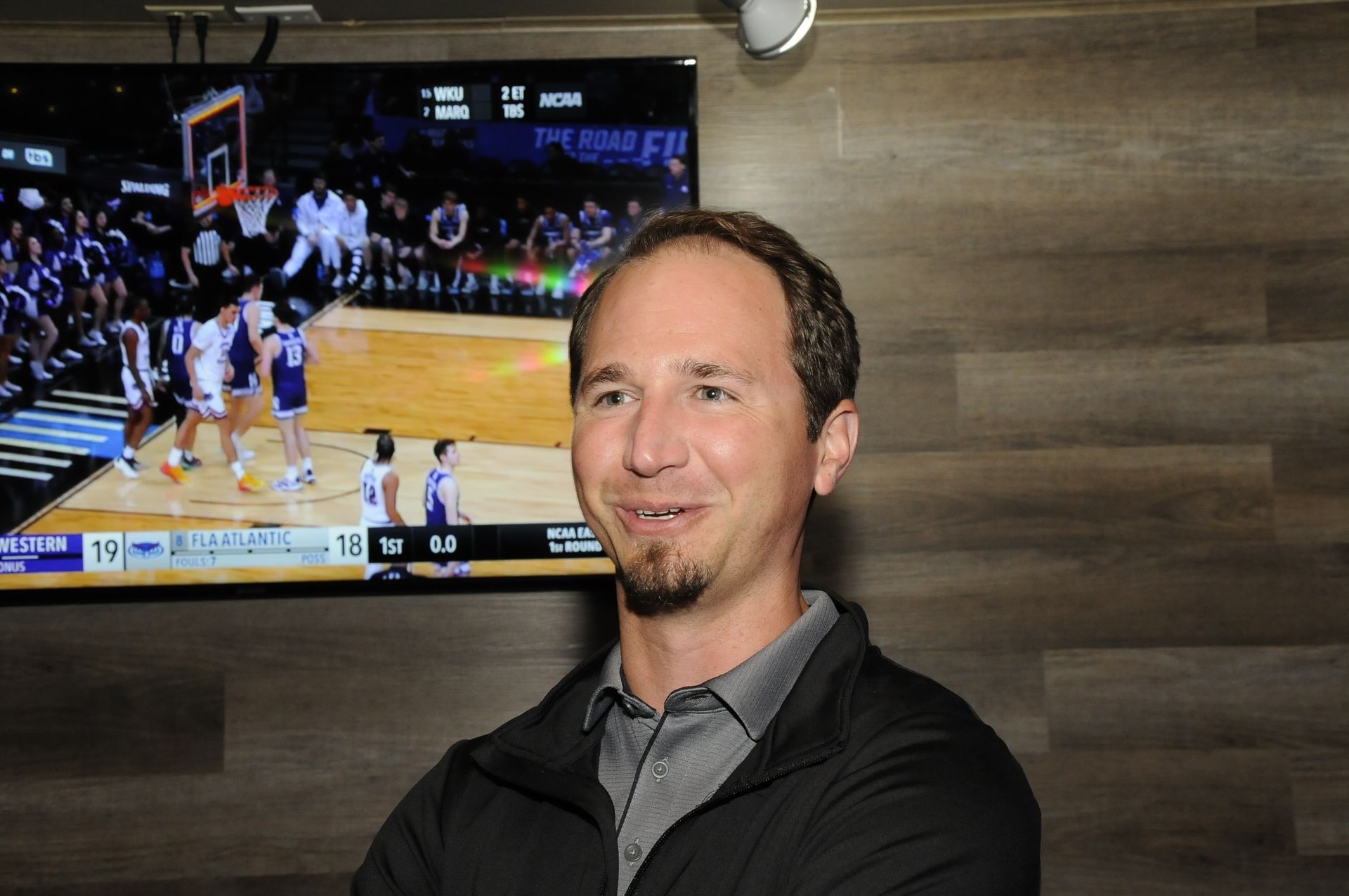 A man is standing in front of a basketball game on a television.