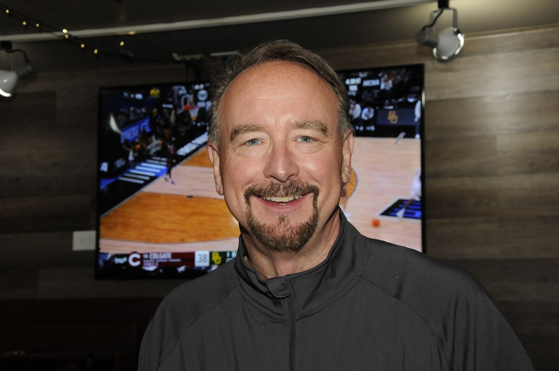 A man with a beard is smiling in front of a basketball screen