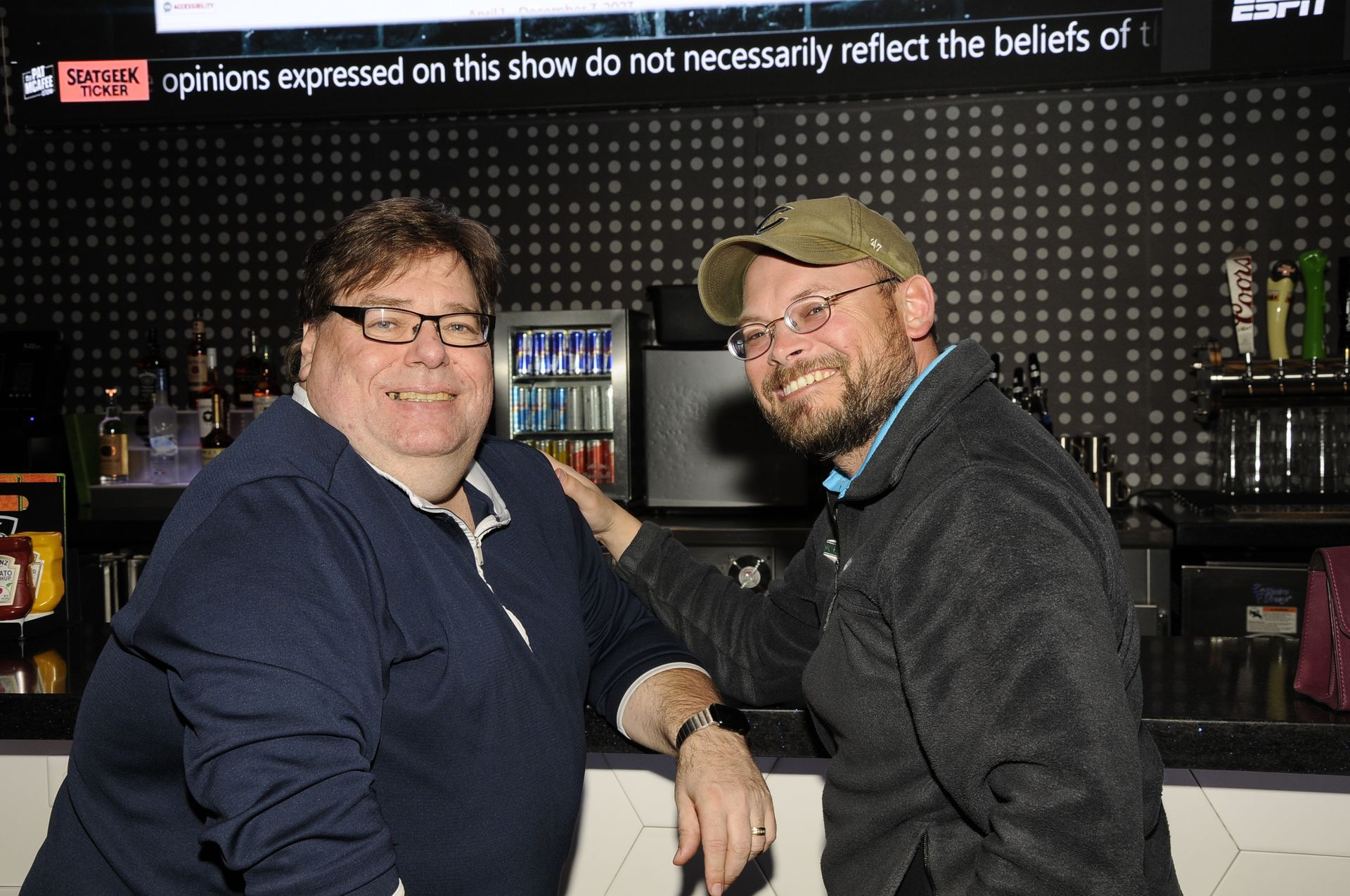 Two men standing next to each other in front of a sign that says opinions expressed on this show do not necessarily reflect the beliefs of