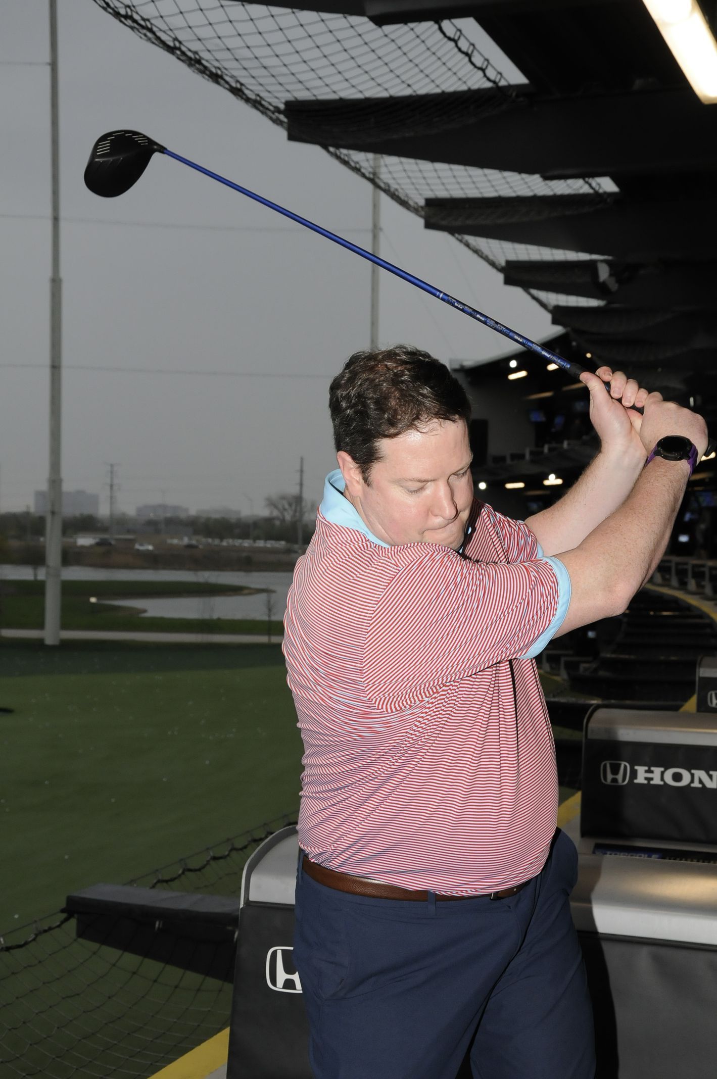 A man in a red and white shirt is swinging a golf club