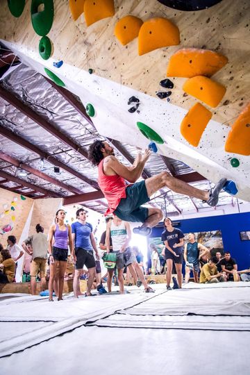 Man bouldering, reaching for a hold on an angled wall. Crowd of people watches in an indoor climbing gym.