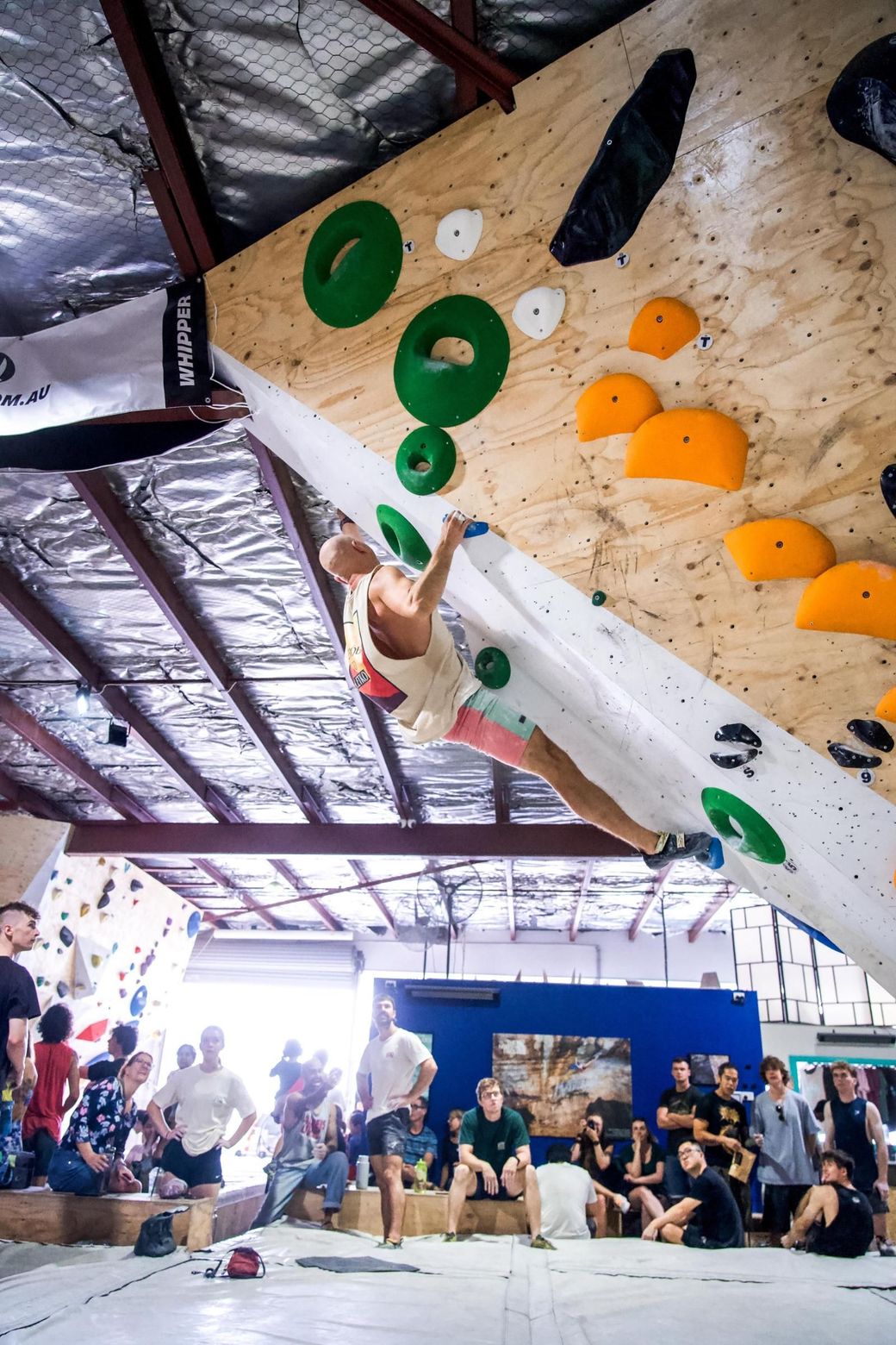 Person climbing an indoor rock wall with onlookers below. Brightly colored holds on wood.