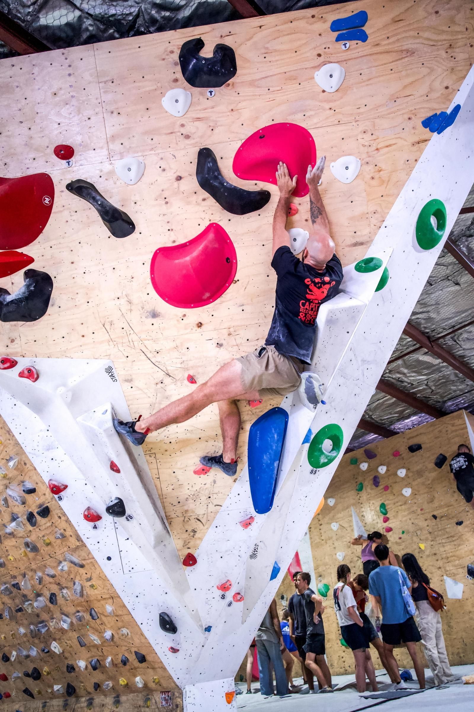Rock climber reaching for a large red hold on a textured wall indoors, people watching.