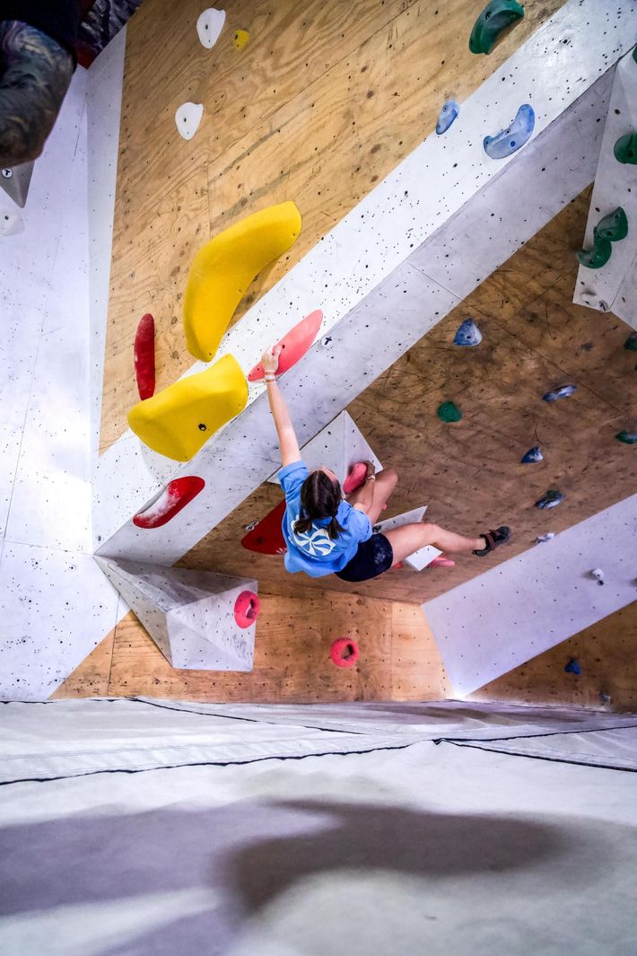 Rock climber in blue shirt reaches for yellow hold on angled climbing wall.