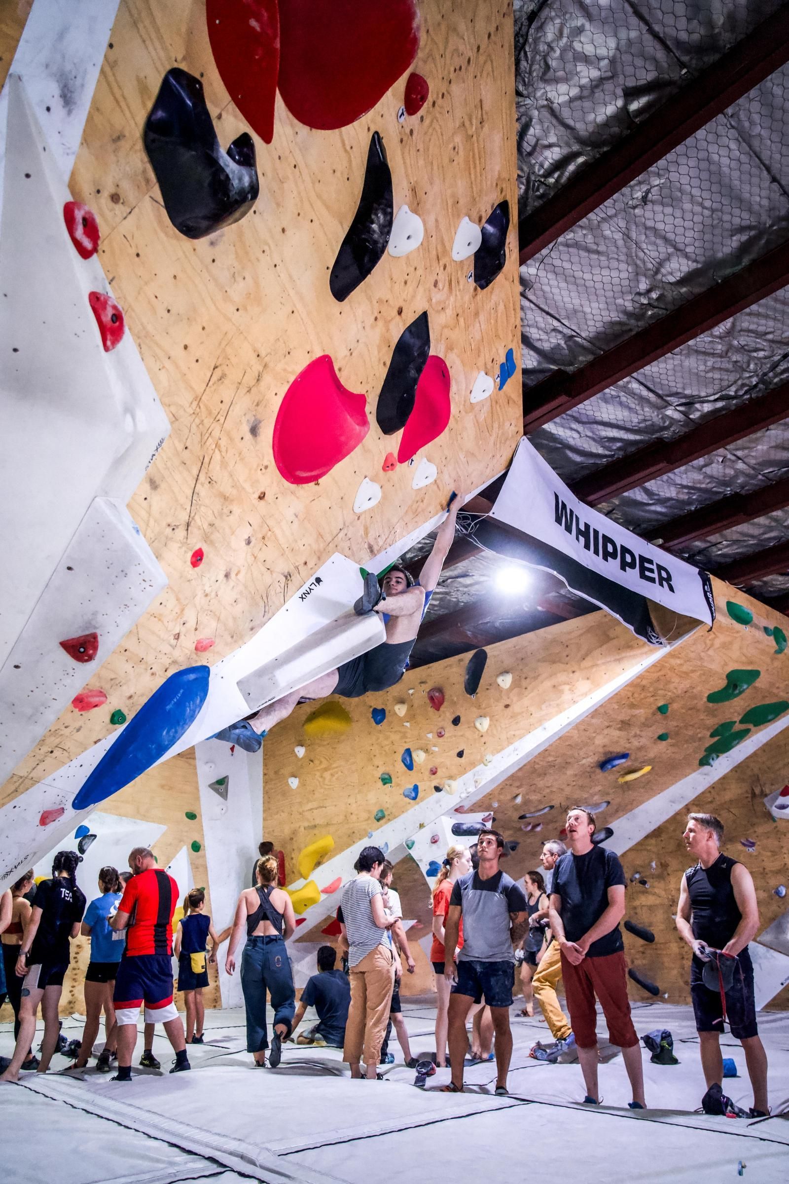 Person climbing an indoor rock wall with onlookers below. Wall has colorful holds. 